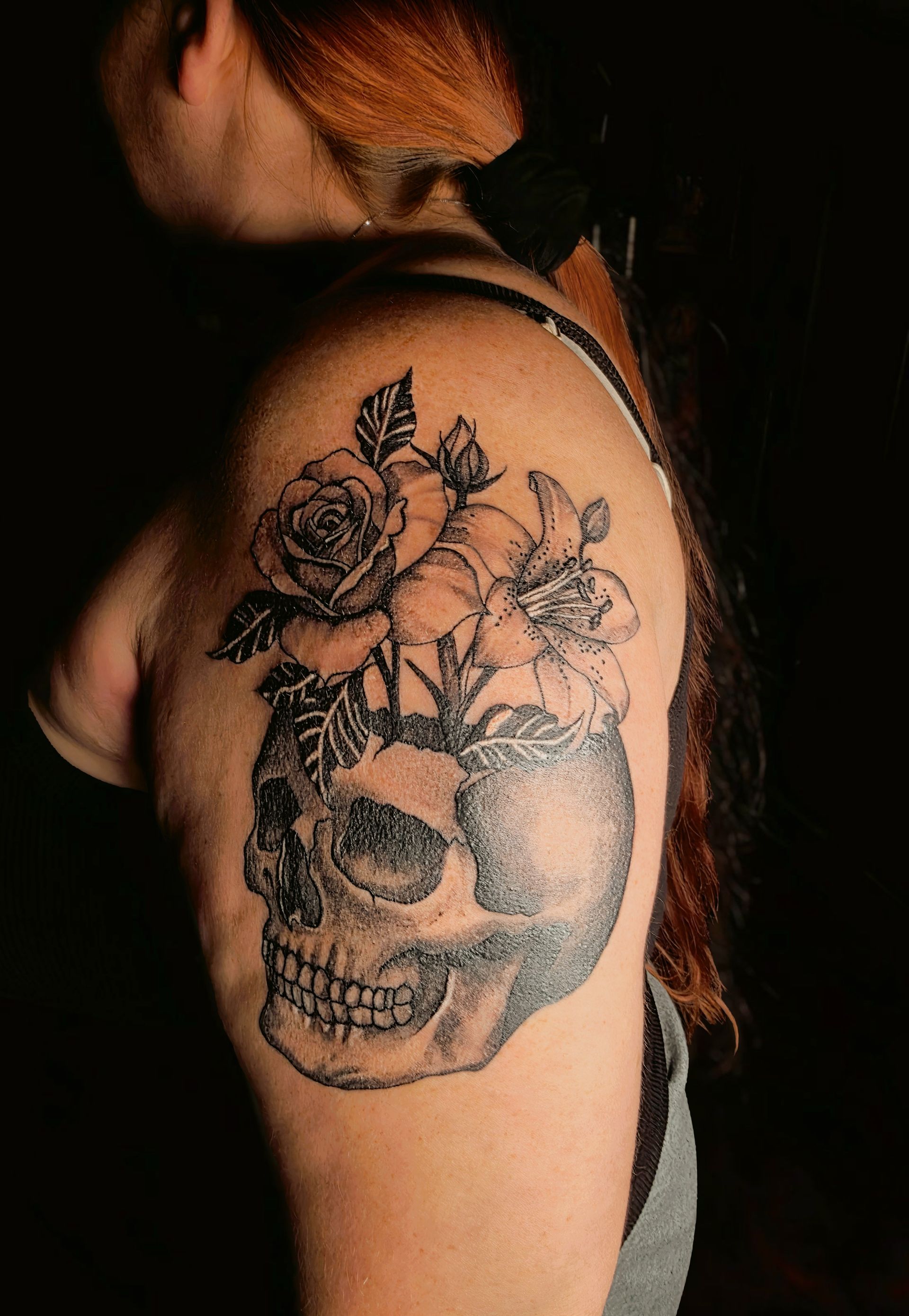 Tattoo of a skull with flowers blossoming from its head, on a person's shoulder.
