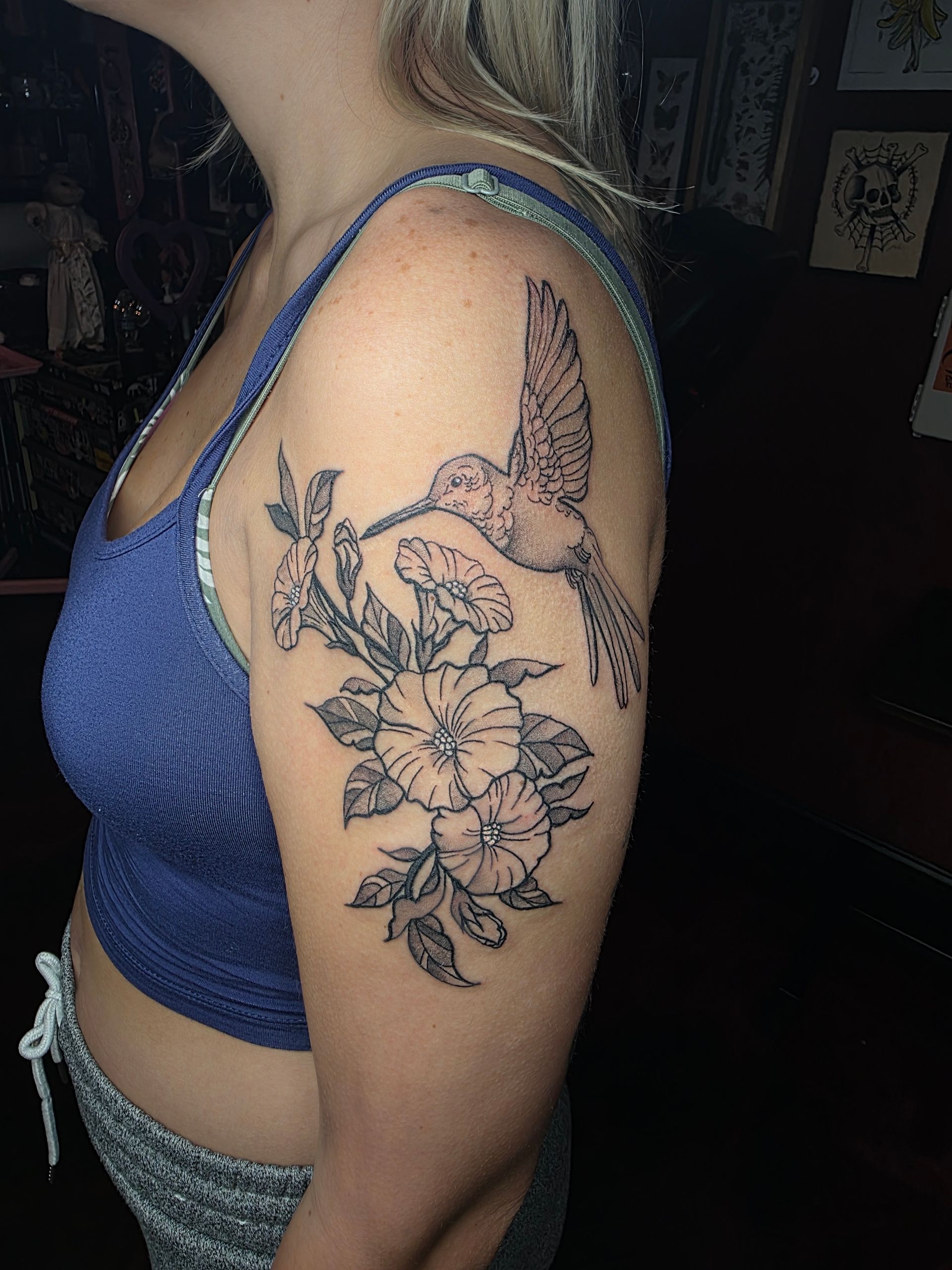 Woman's arm with black ink tattoo of hummingbird and flowers. Blue tank top, dark background.