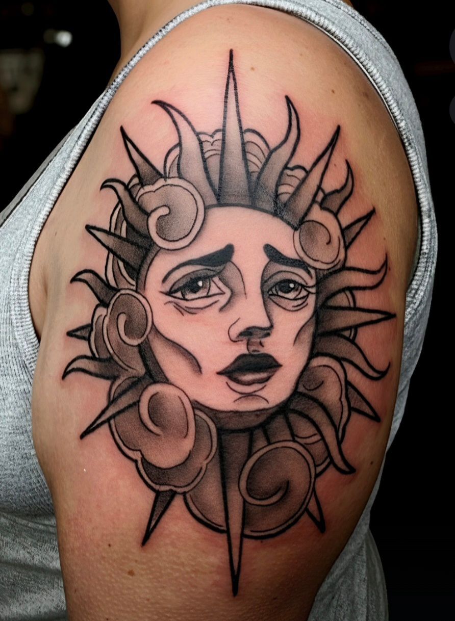 Sun face tattoo on a person's upper arm, featuring rays and swirling clouds; grayscale.