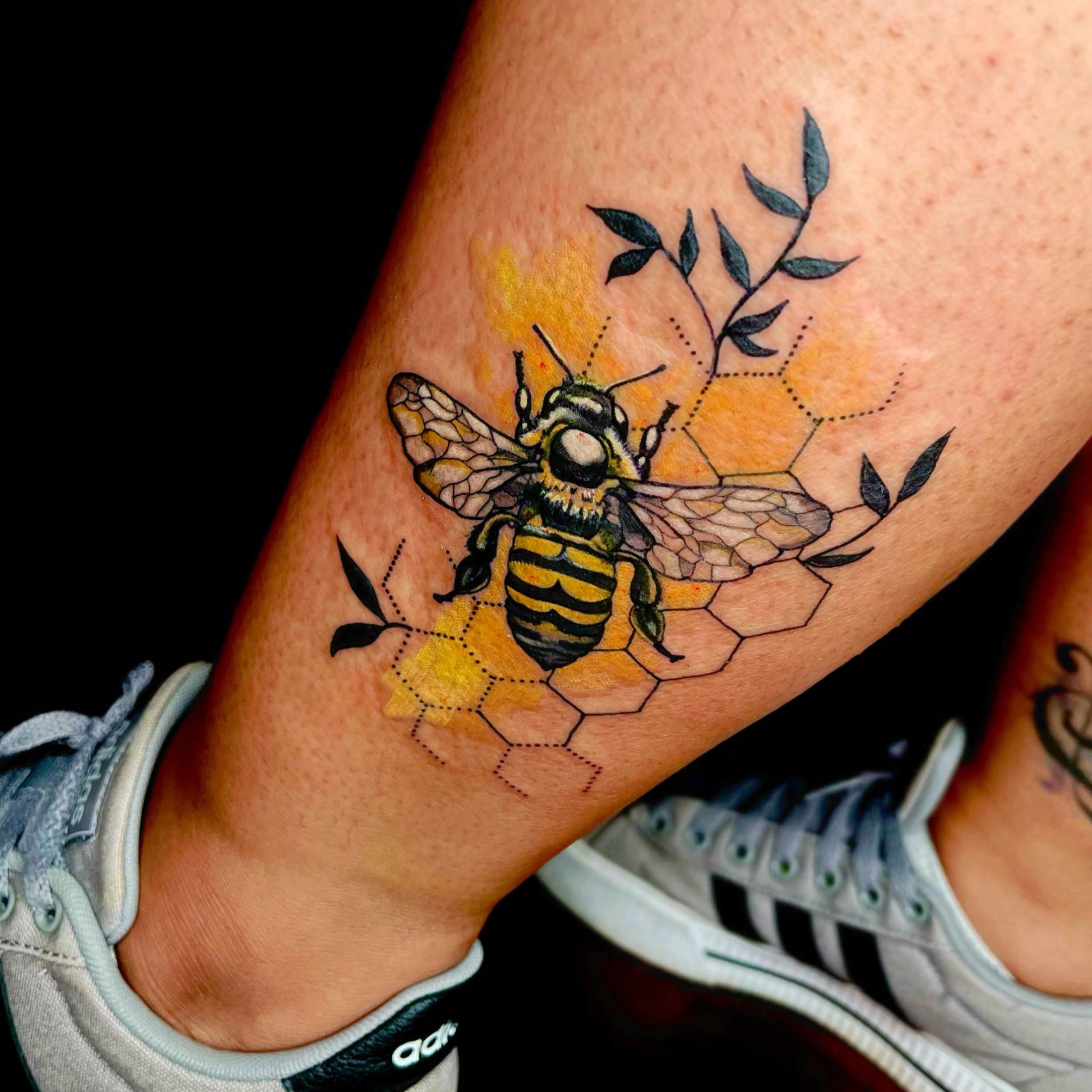 Bee tattoo with yellow honeycomb and leaves on leg, wearing grey Adidas sneaker.