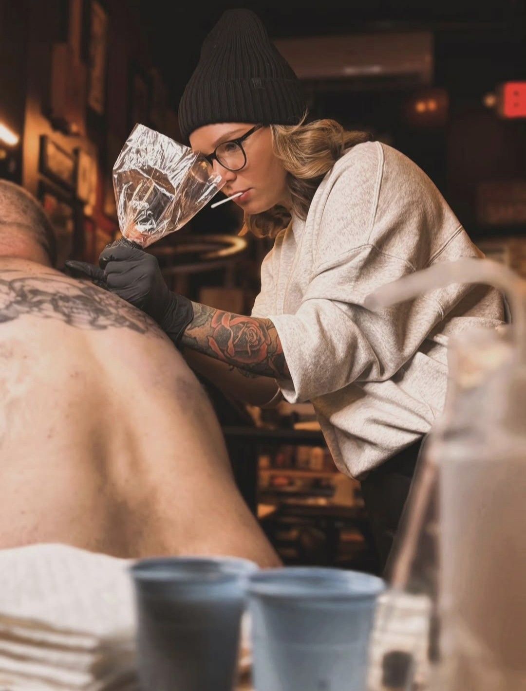 A person with tattoos, wearing a beanie and glasses, tattoos a person's back in a dimly lit setting.