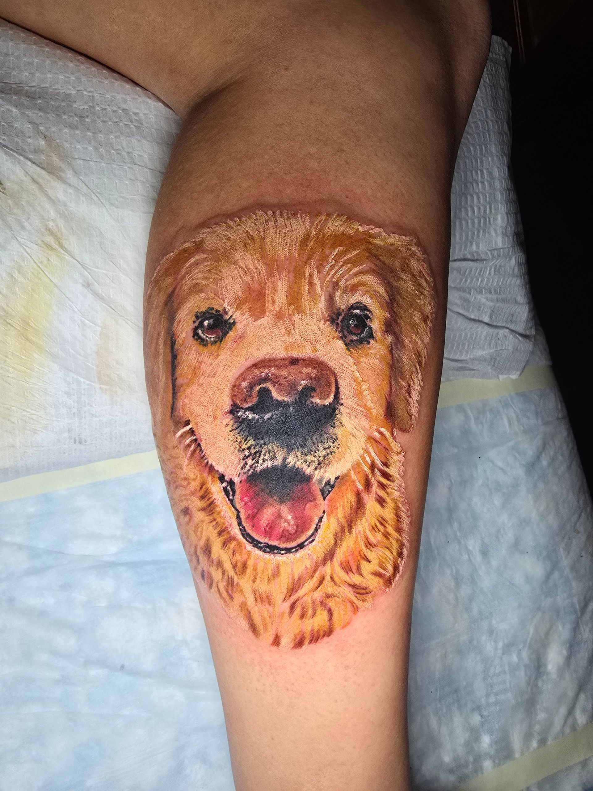 Golden retriever dog face tattoo on a person's arm, showcasing a smiling expression with a pink tongue.