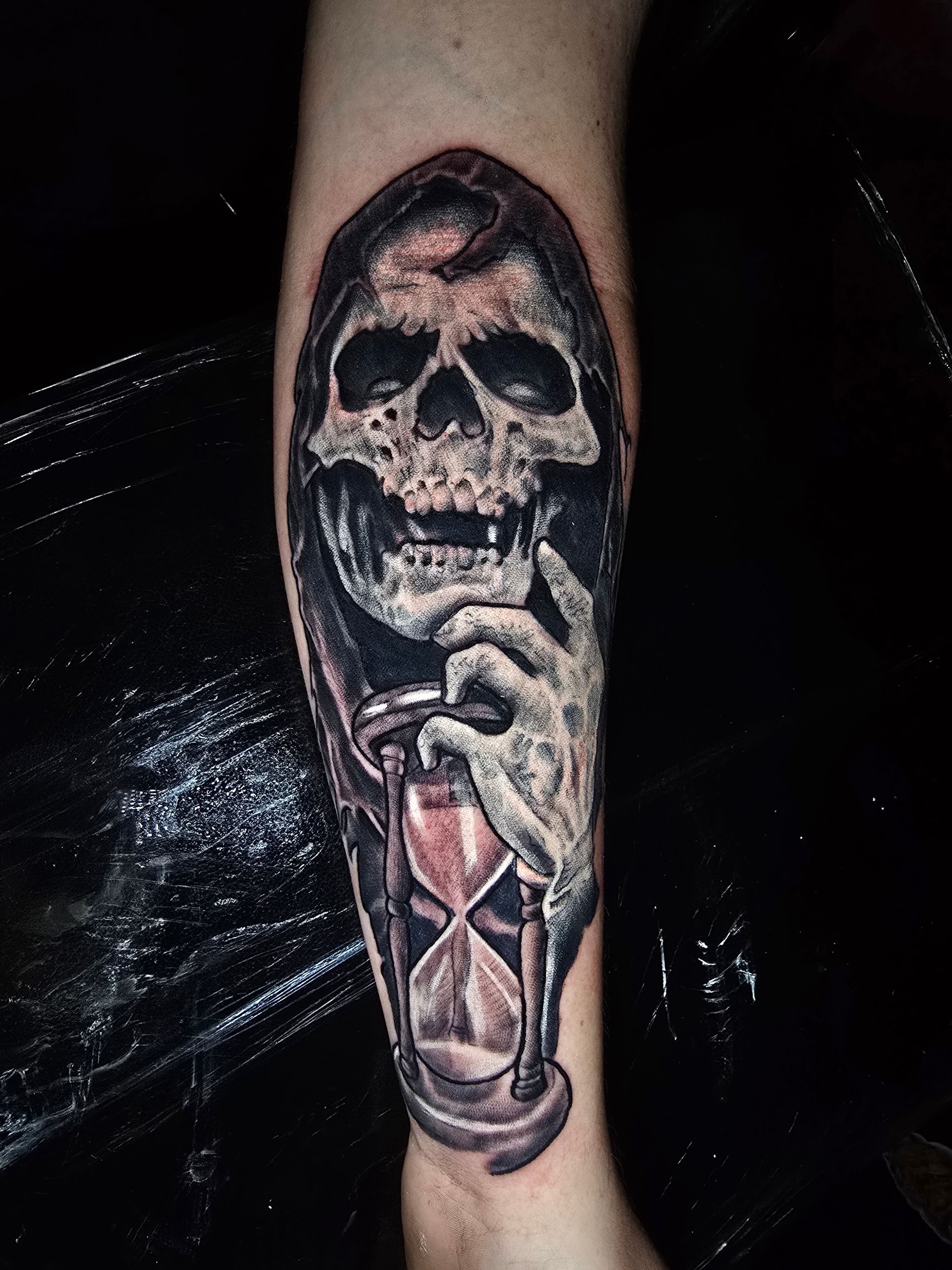 Tattoo of a skull figure holding an hourglass, with a black cloak and shading.