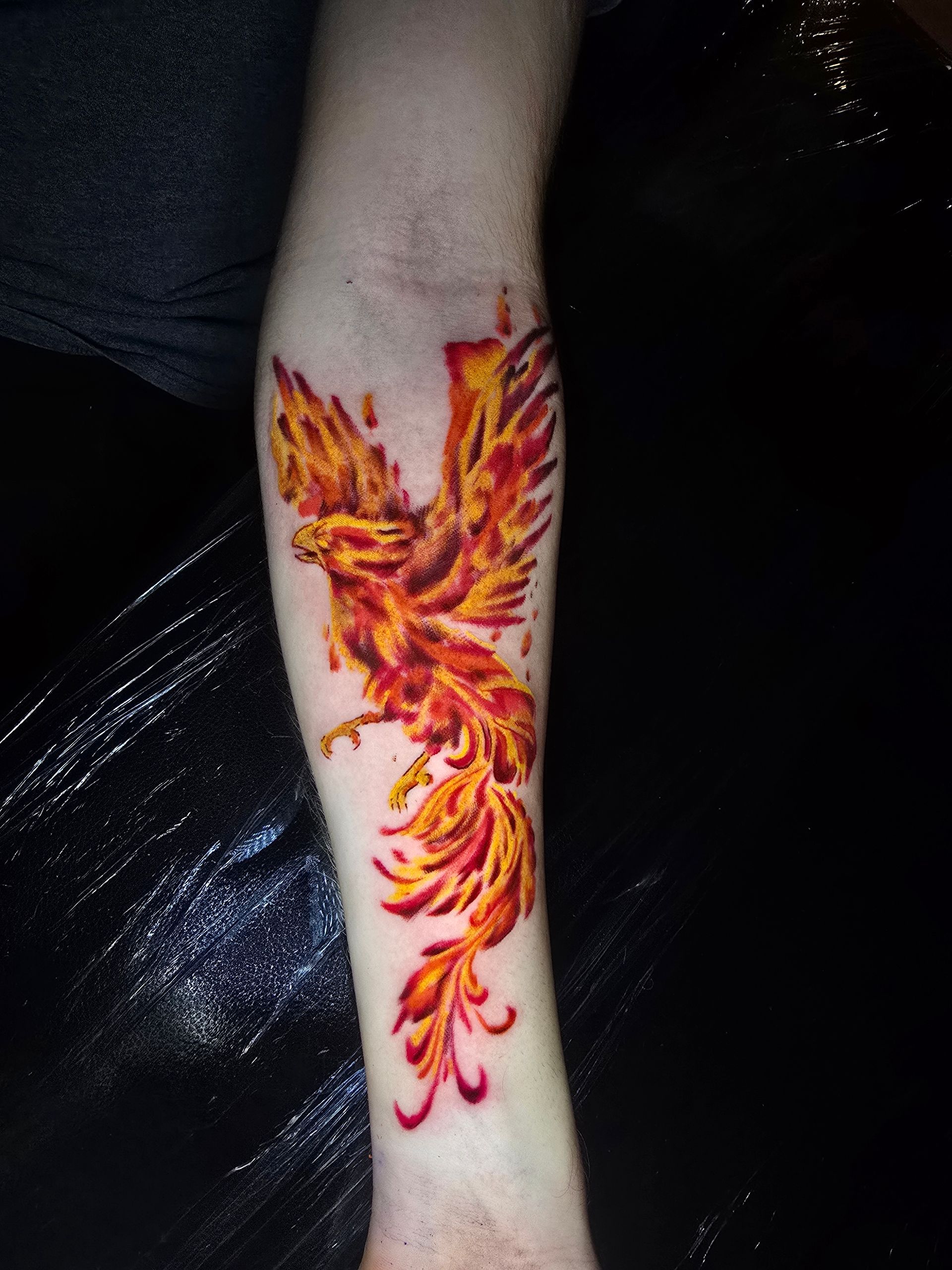 Tattoo of a fiery phoenix rising, with orange and yellow flames, on a forearm.