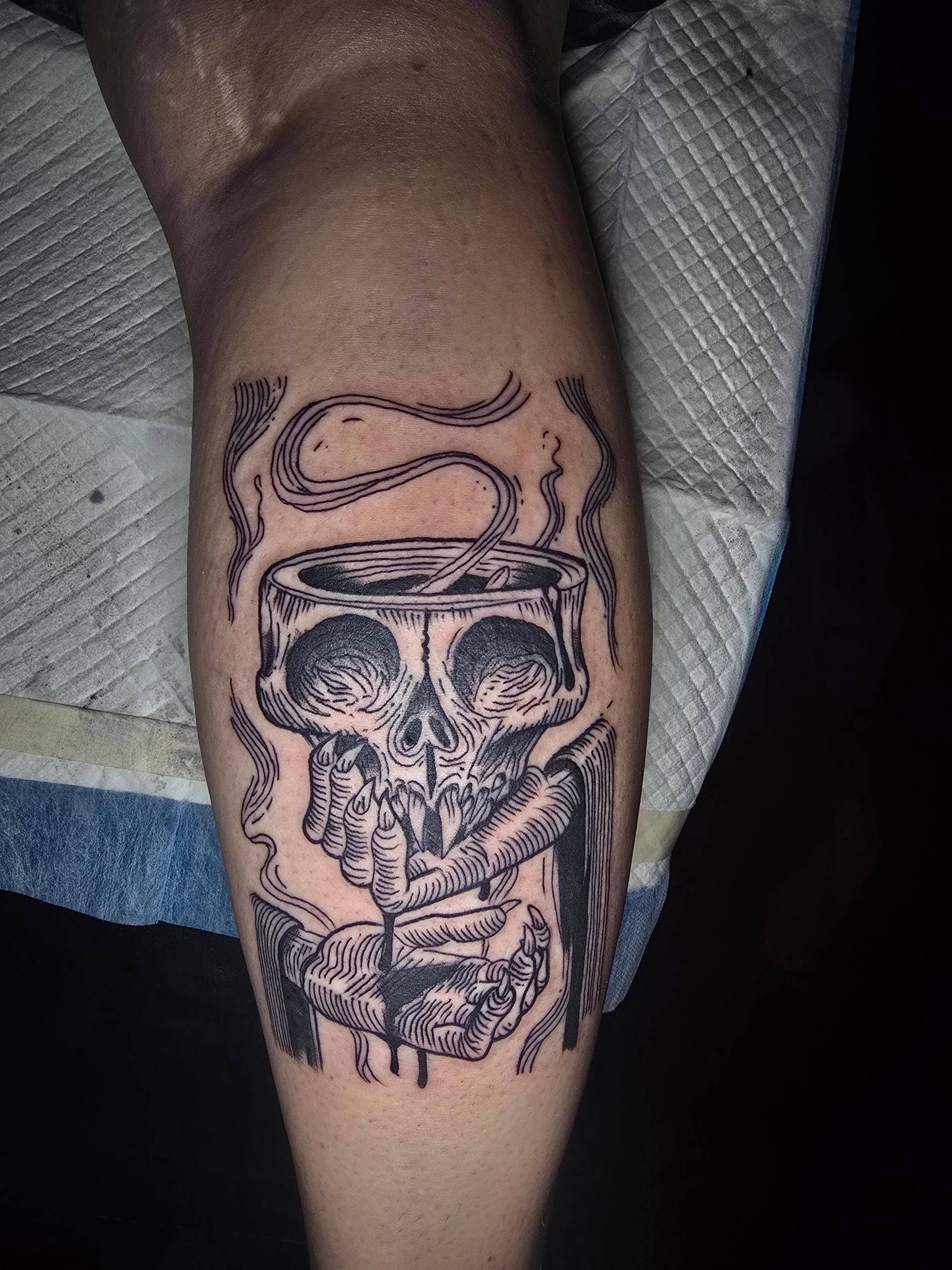 Tattoo of a skull and coffee cup on a leg. Black ink, gray shading, with smoke effect.