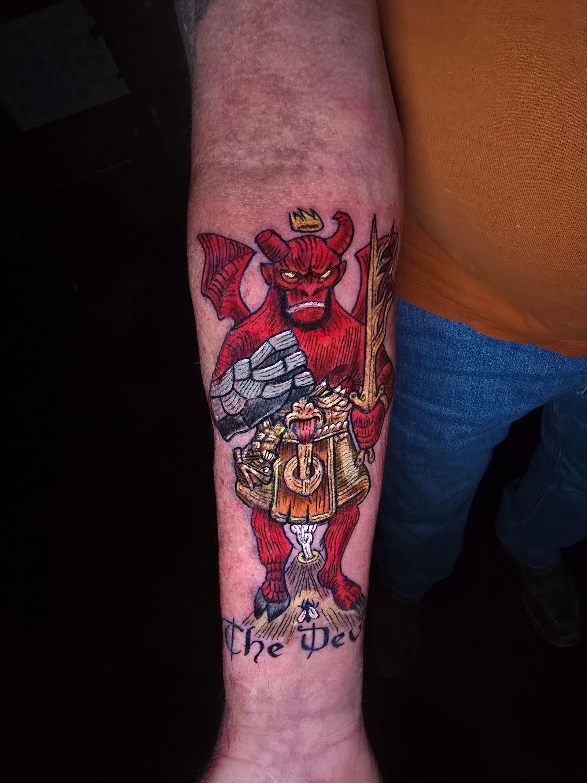 Tattoo of a red devil with a crown, holding a sword and wearing a kilt, on a person's arm, with text 