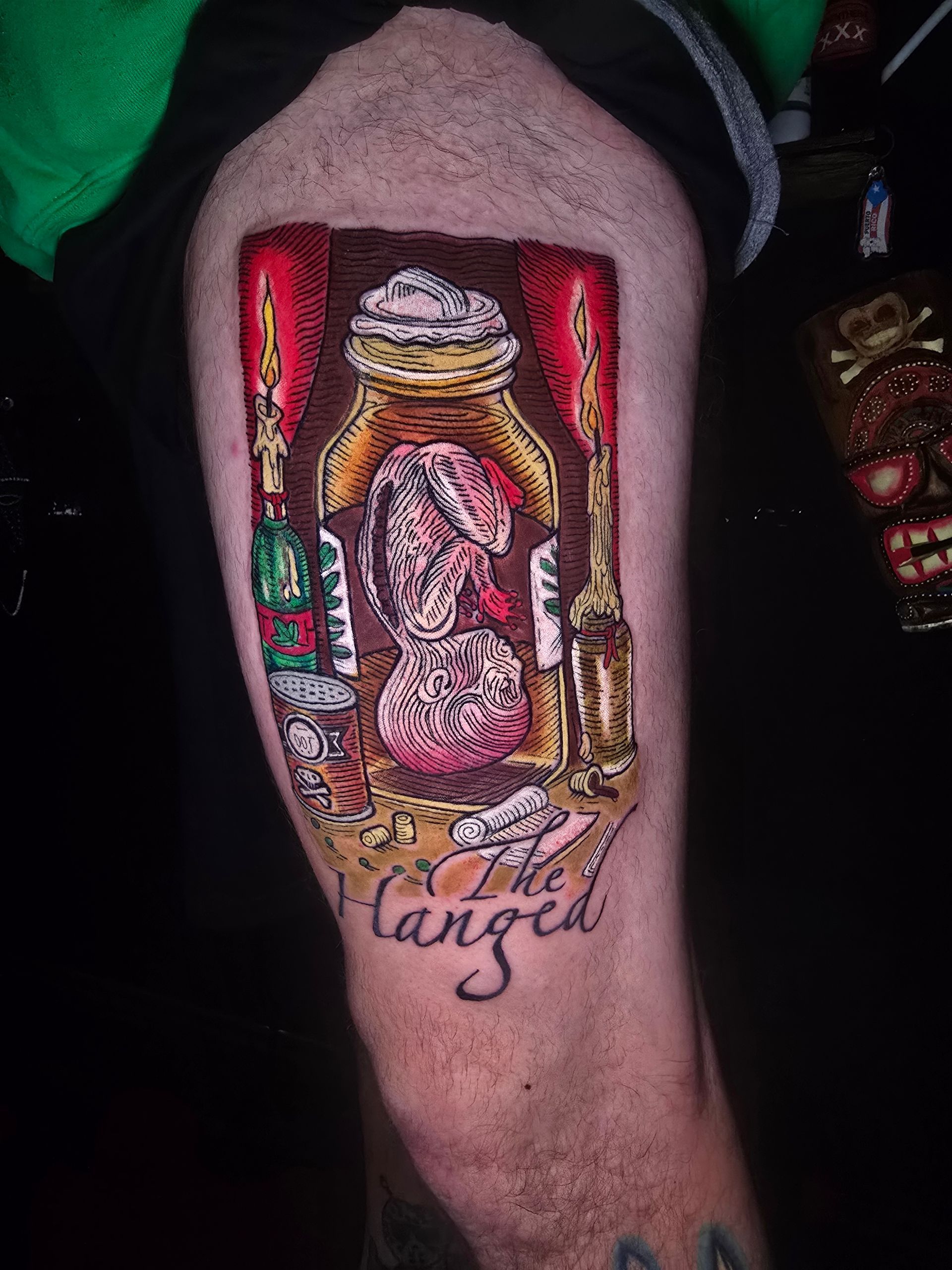 Tattoo on leg: jar with brain-like objects, candles, and “The Hanged.” Red, gold, and brown colors.