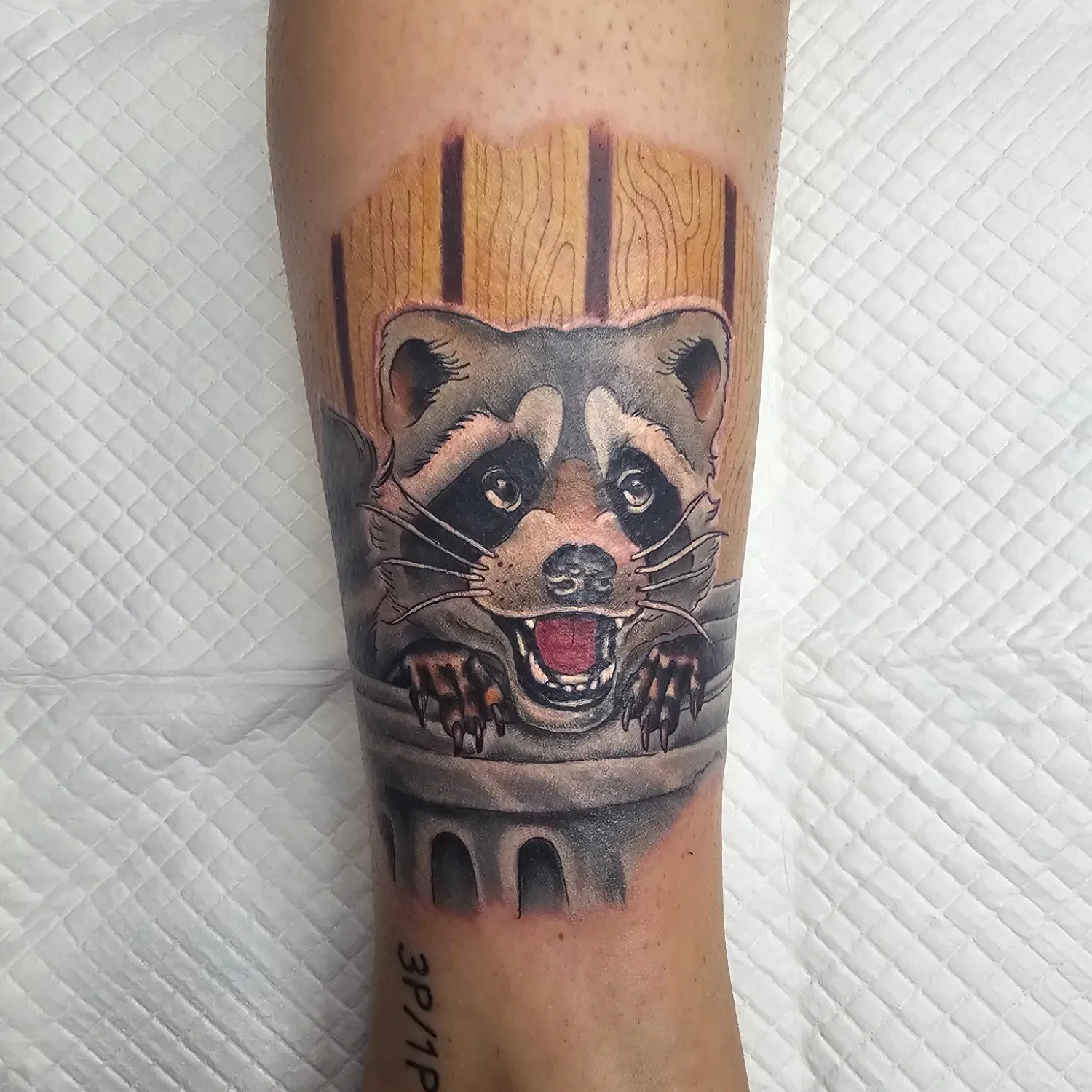 Raccoon tattoo emerging from a wooden barrel. The raccoon is smiling with red mouth and has gray, brown, and black fur.