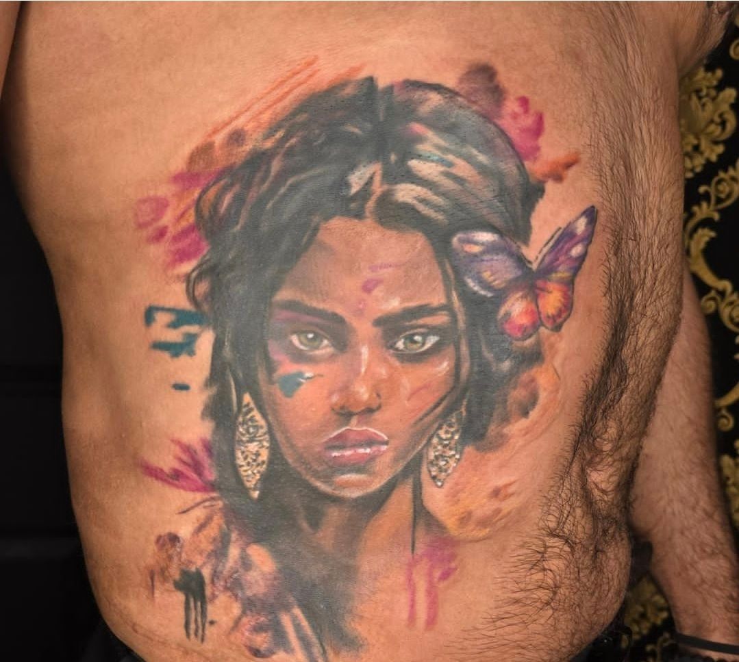 Tattoo of a woman's face with a butterfly. Watercolor style, colorful accents. Centered on a torso.
