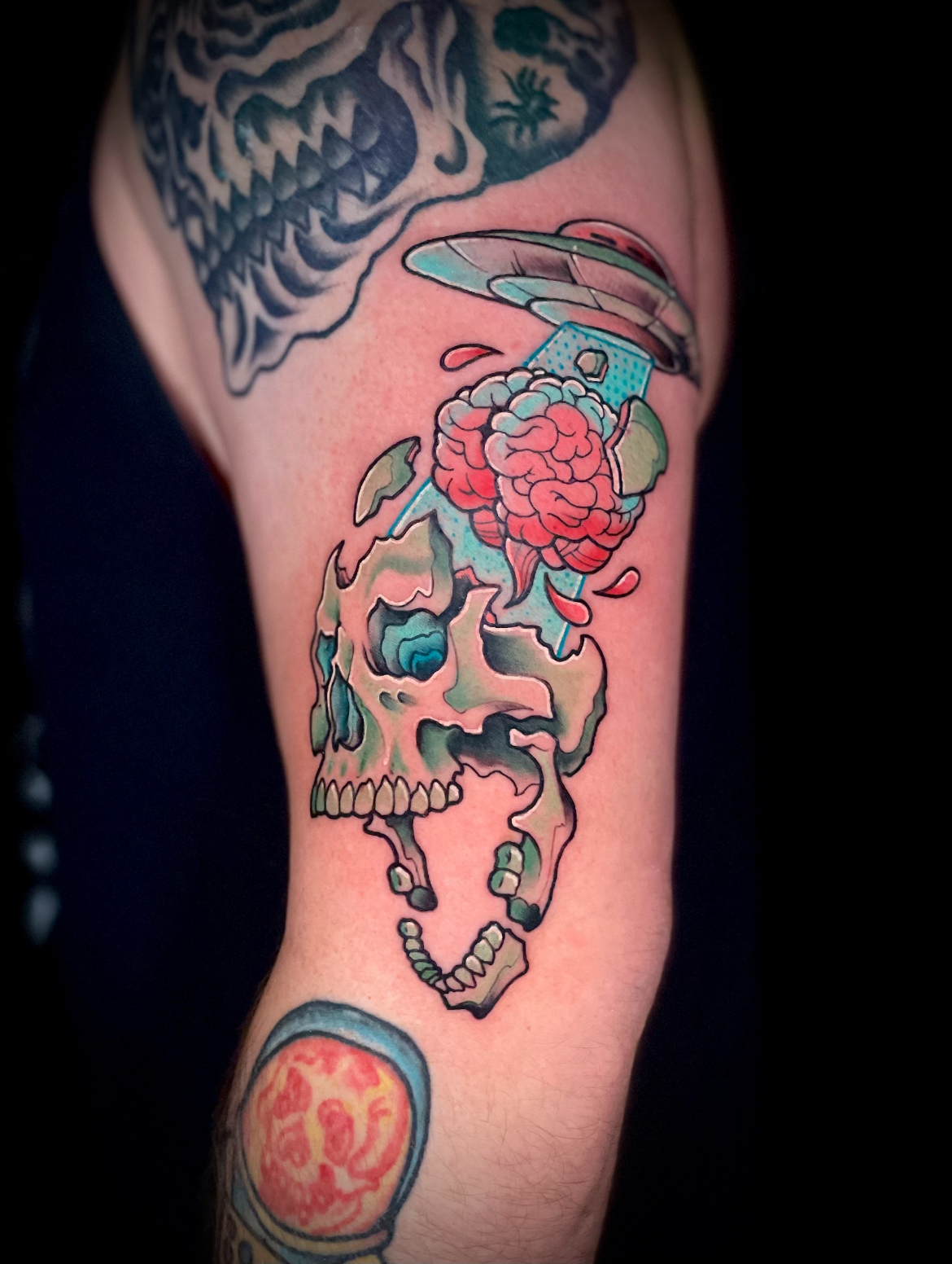 Tattoo of a skull being abducted by a UFO, showing exposed brain. Blue, red, green, and tan colors.