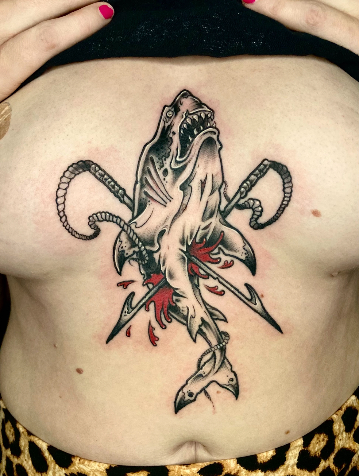 Tattoo of a shark impaled by spears, with red blood detail, on a person's chest.