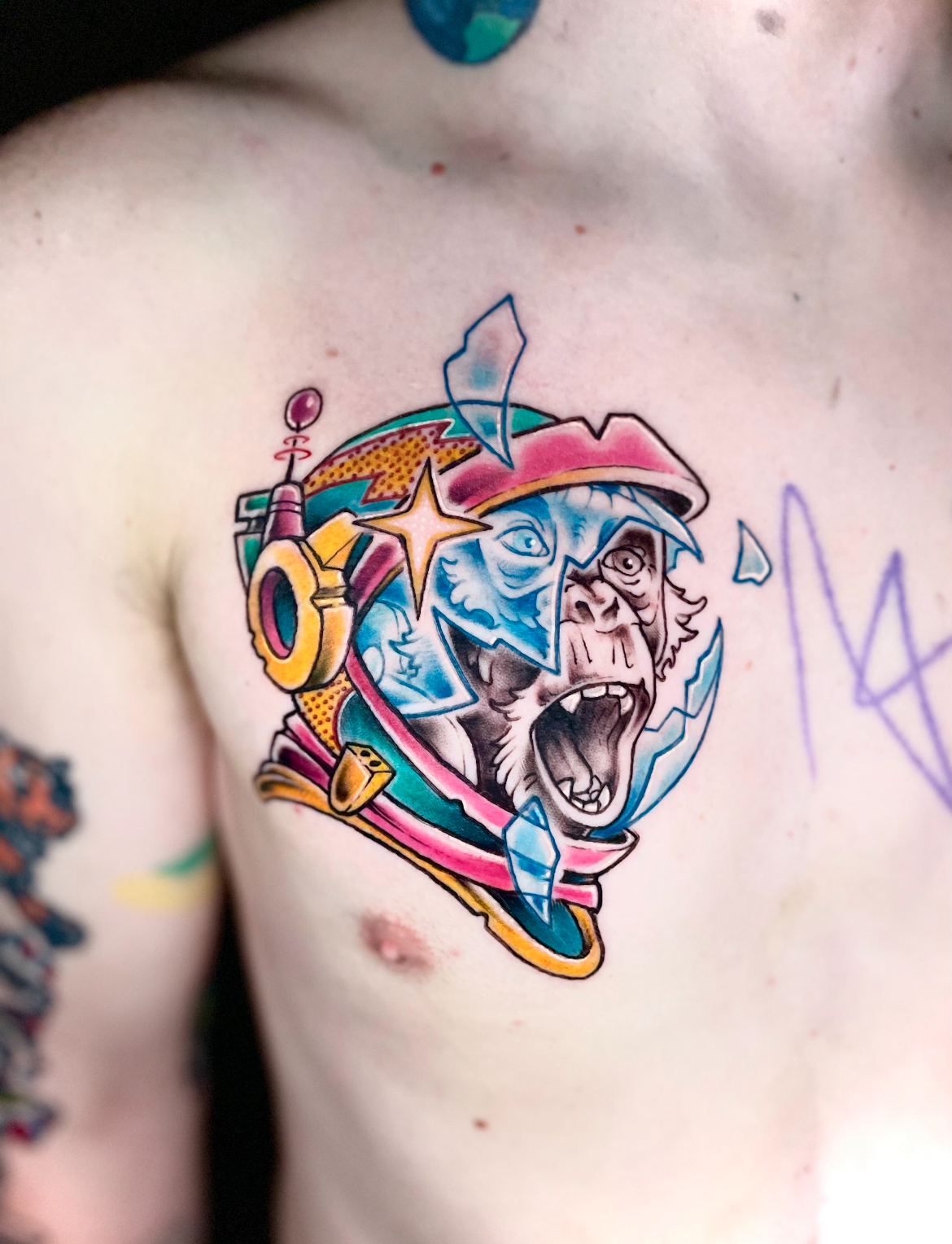 Colorful tattoo on chest: monkey with an open mouth inside a cracked helmet, surrounded by planet-like shapes.