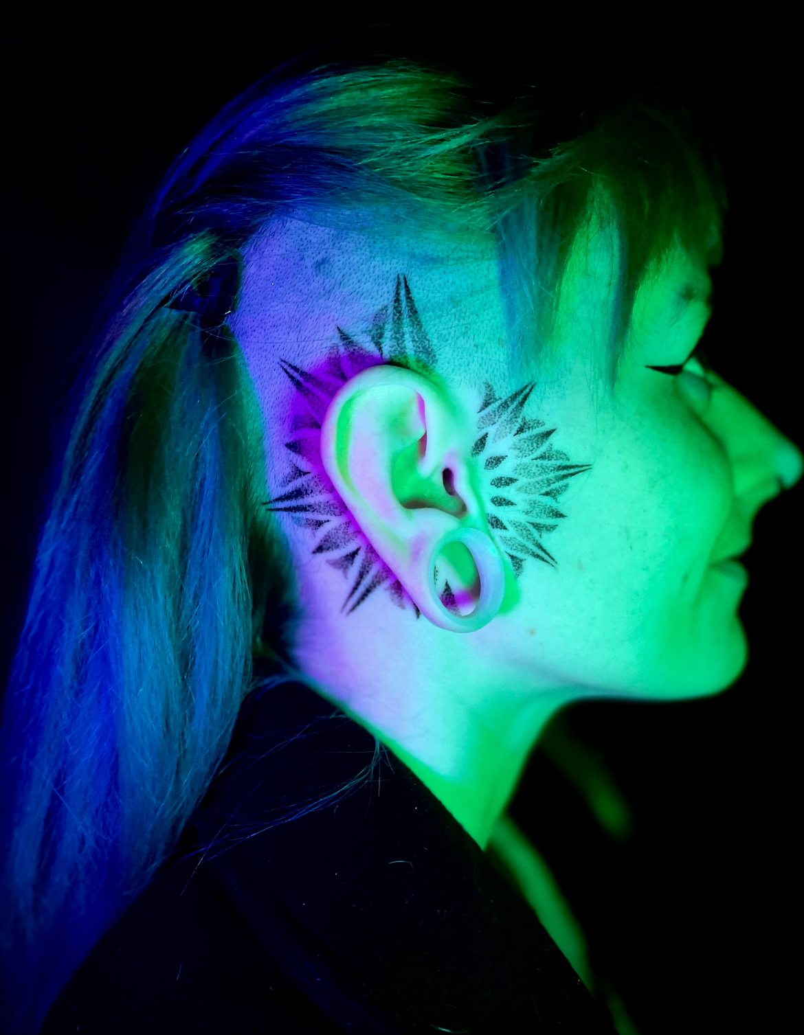 Woman with ear tattoo, side profile. Green and blue lighting. Spiked design around ear and shaved side of head.