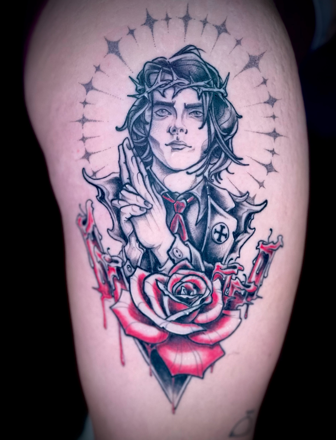 Tattoo of a person with a crown of thorns, halo, and clasped hands. A red rose and a dripping tattoo gun are below.