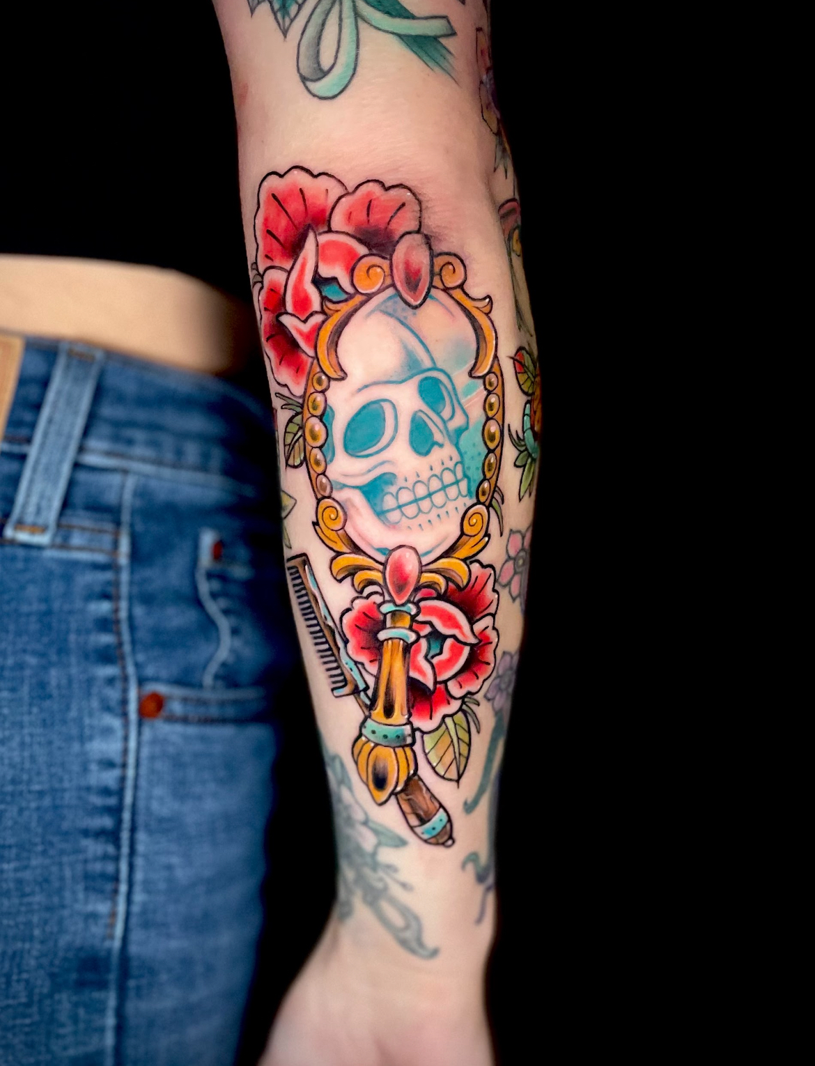 Tattoo on forearm: skull in a golden mirror, red flowers. Blue, gold, white, and black ink.