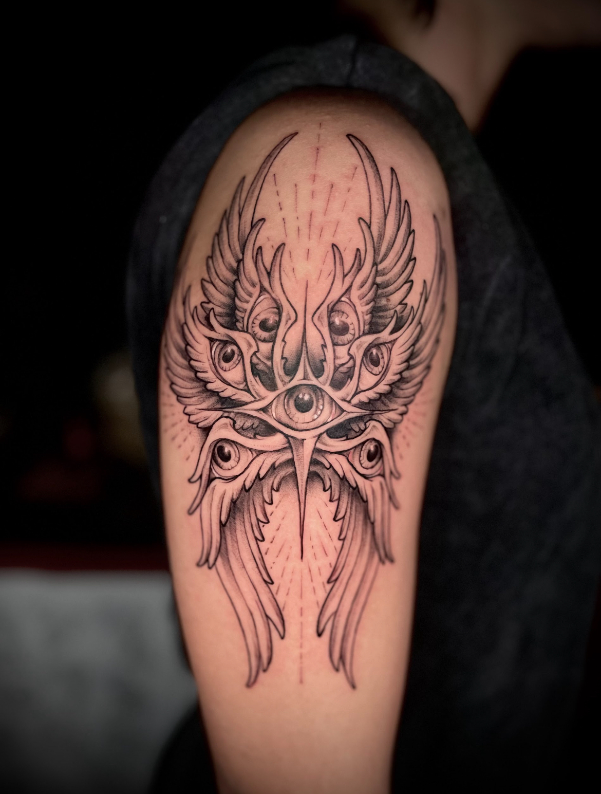 Tattoo of an angel-like figure with multiple eyes and wings on a person's upper arm.