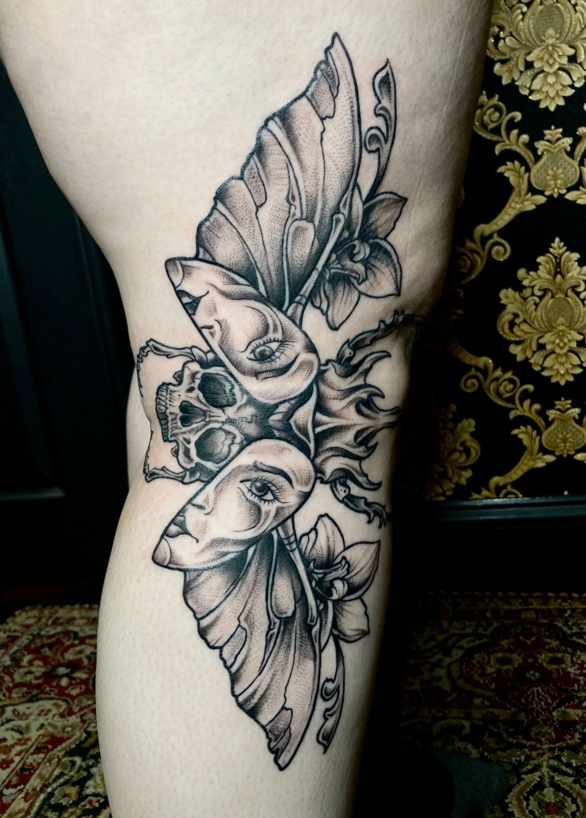 Tattoo on a leg featuring a skull, two faces, large butterfly wings, and flowers in black ink.