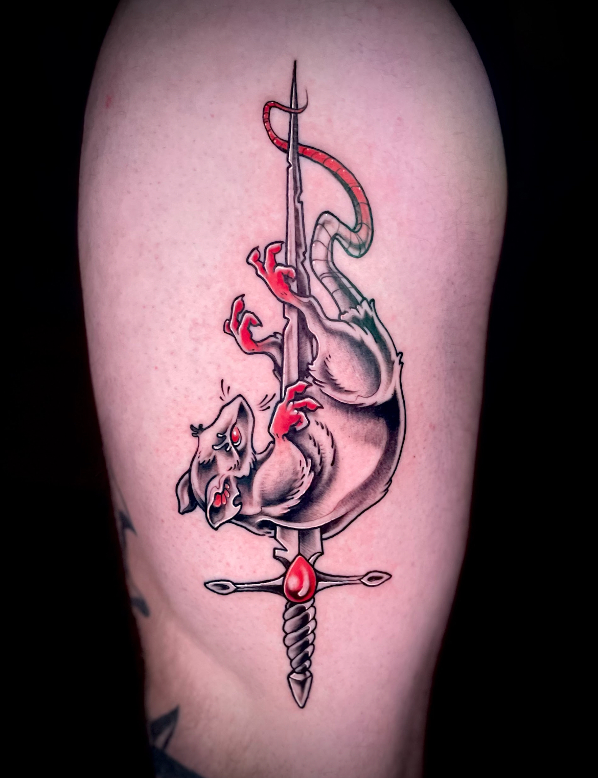 Tattoo of a rat impaled on a sword. The rat is gray with red hands and a tail, and the sword has a red jewel.