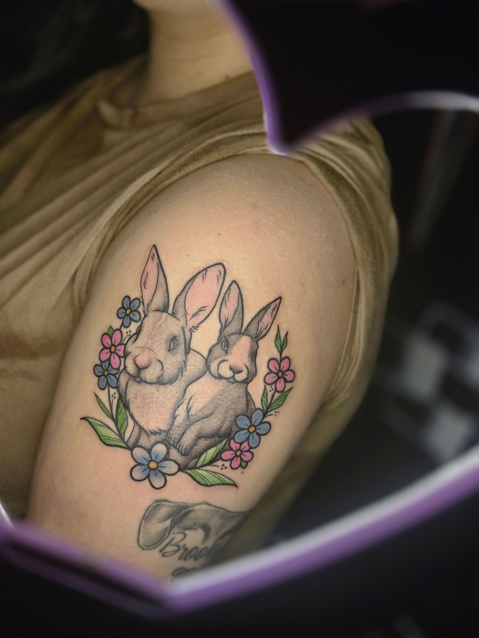 Tattoo of two rabbits in a flower wreath on an arm. Gray and pink tones, soft details.