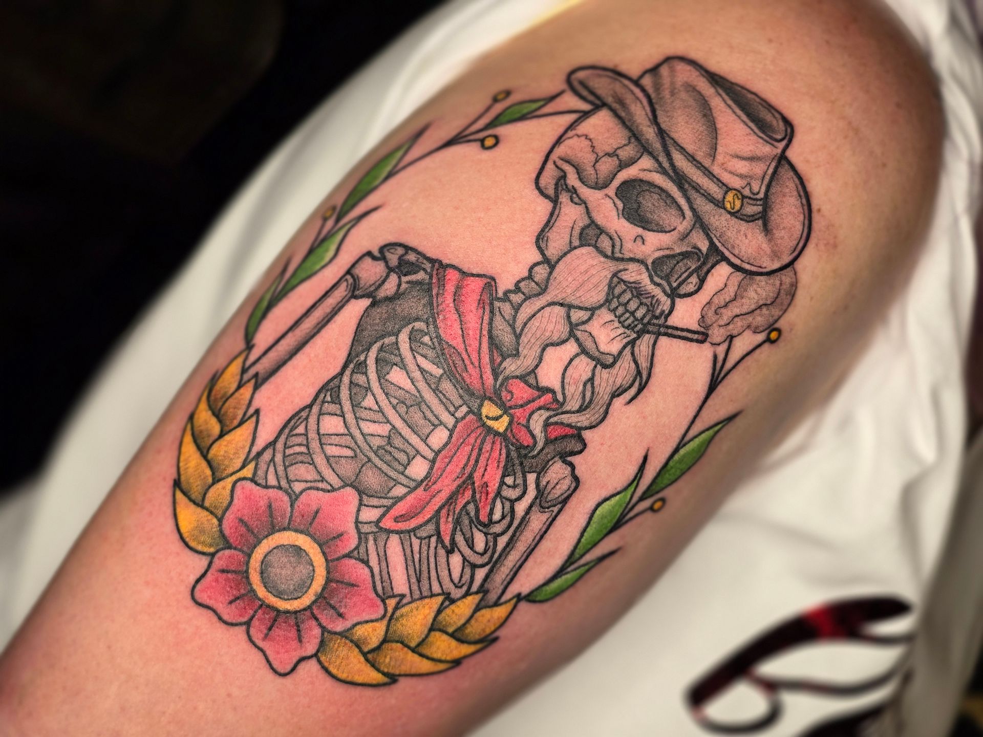 Tattoo of a cowboy skeleton with a hat, bandanna, and cigarette, framed by a wreath with a flower.