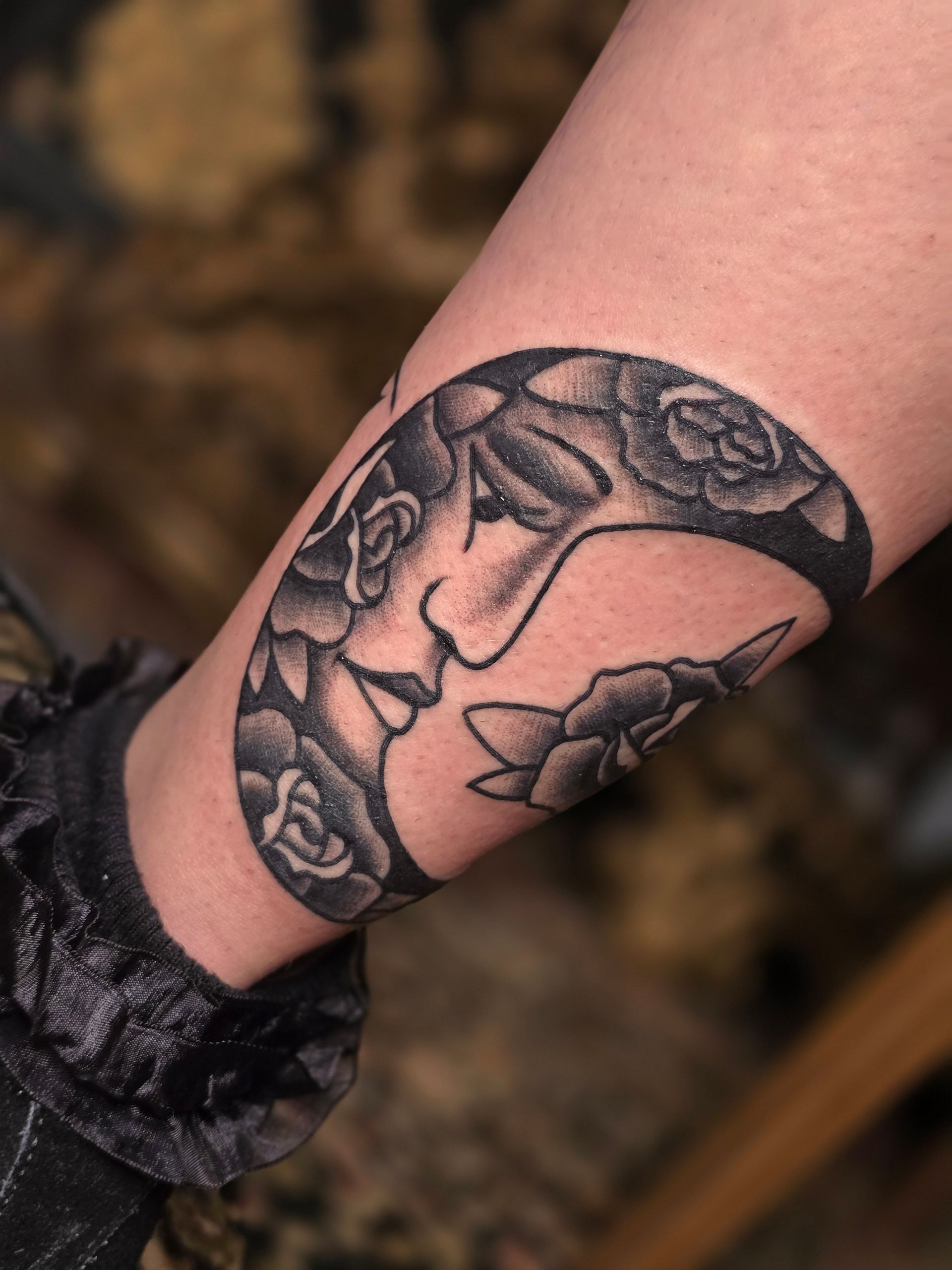 Black ink crescent moon tattoo with a woman's face and roses on a forearm.