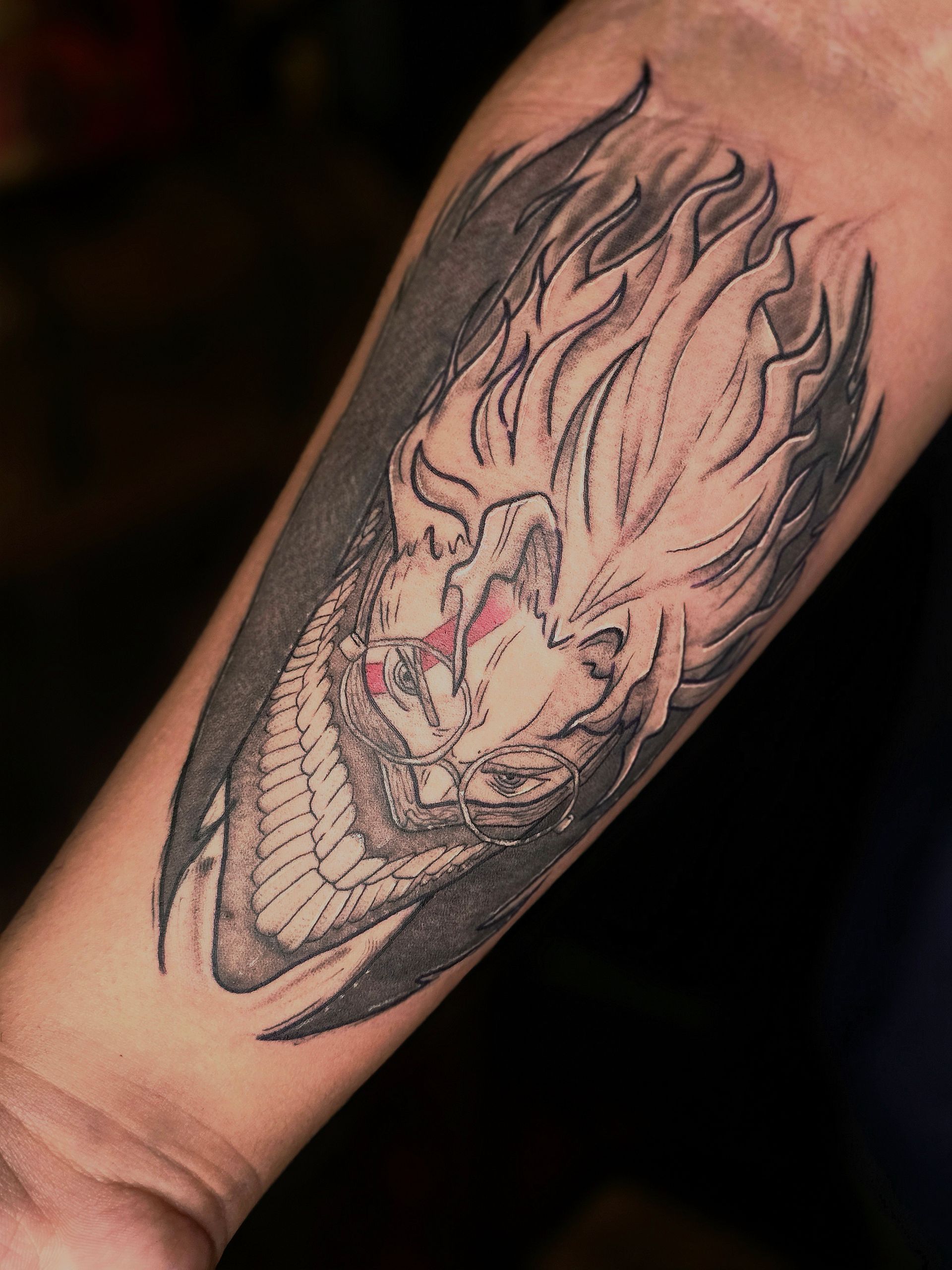 Tattoo on forearm: fierce face with flaming hair, gray shading, red eyes, emerging from dark shape.