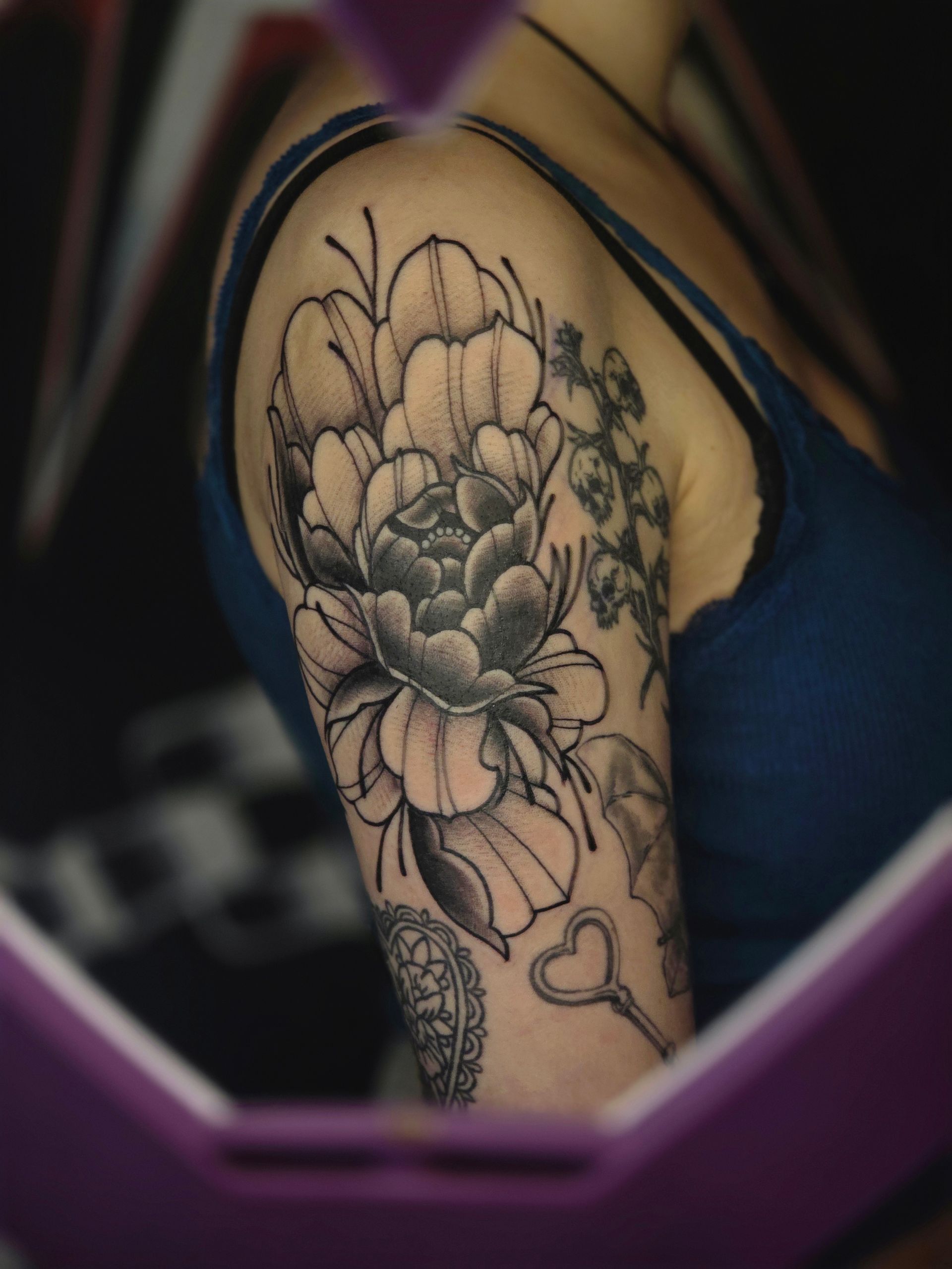 Tattoo of peony and other flowers on a person's upper arm, with key and other designs. Black and white, blue shirt.