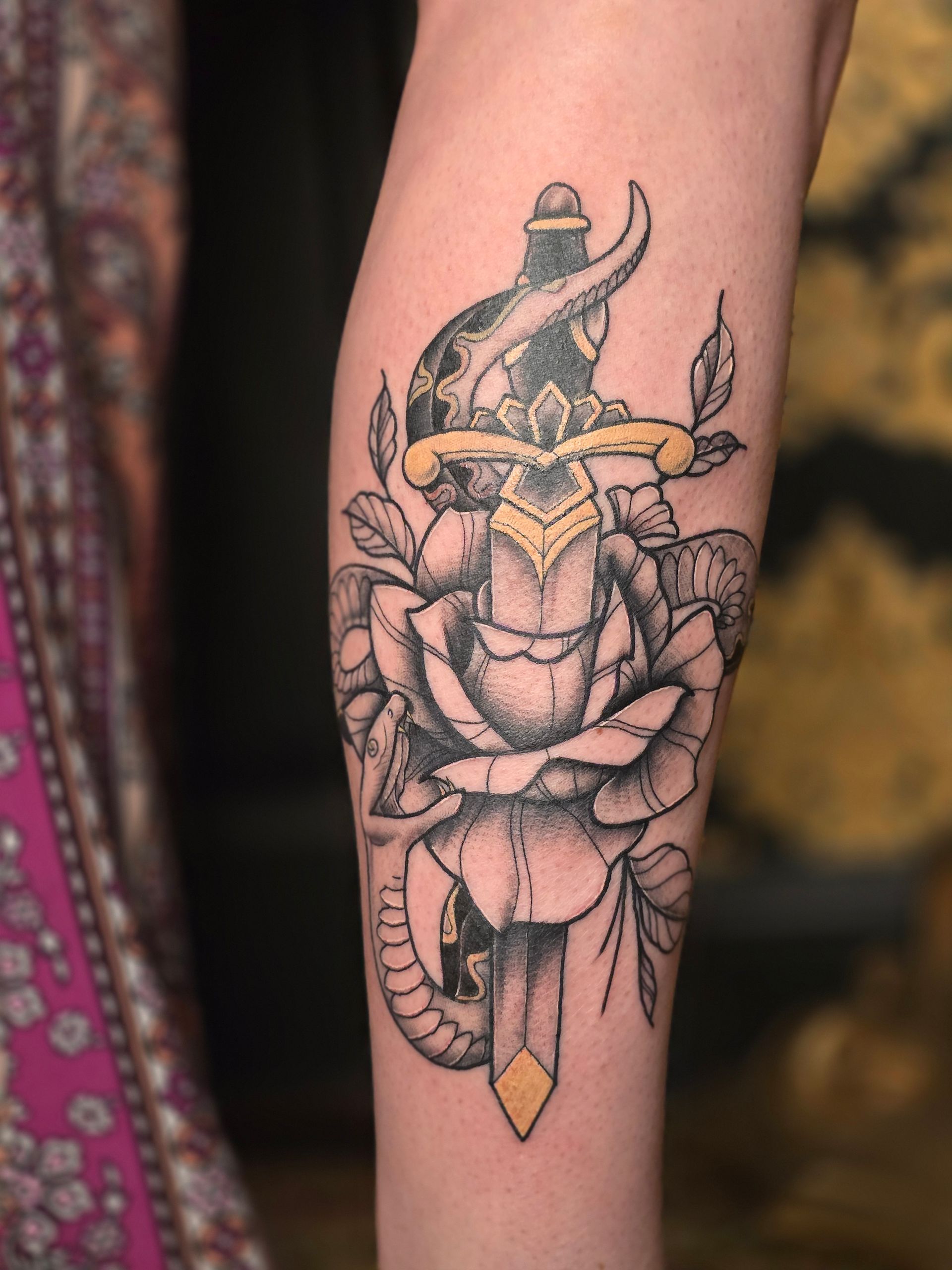 Tattoo of a dagger through a rose on an arm; black, white, and gold colors; floral and weapon elements.