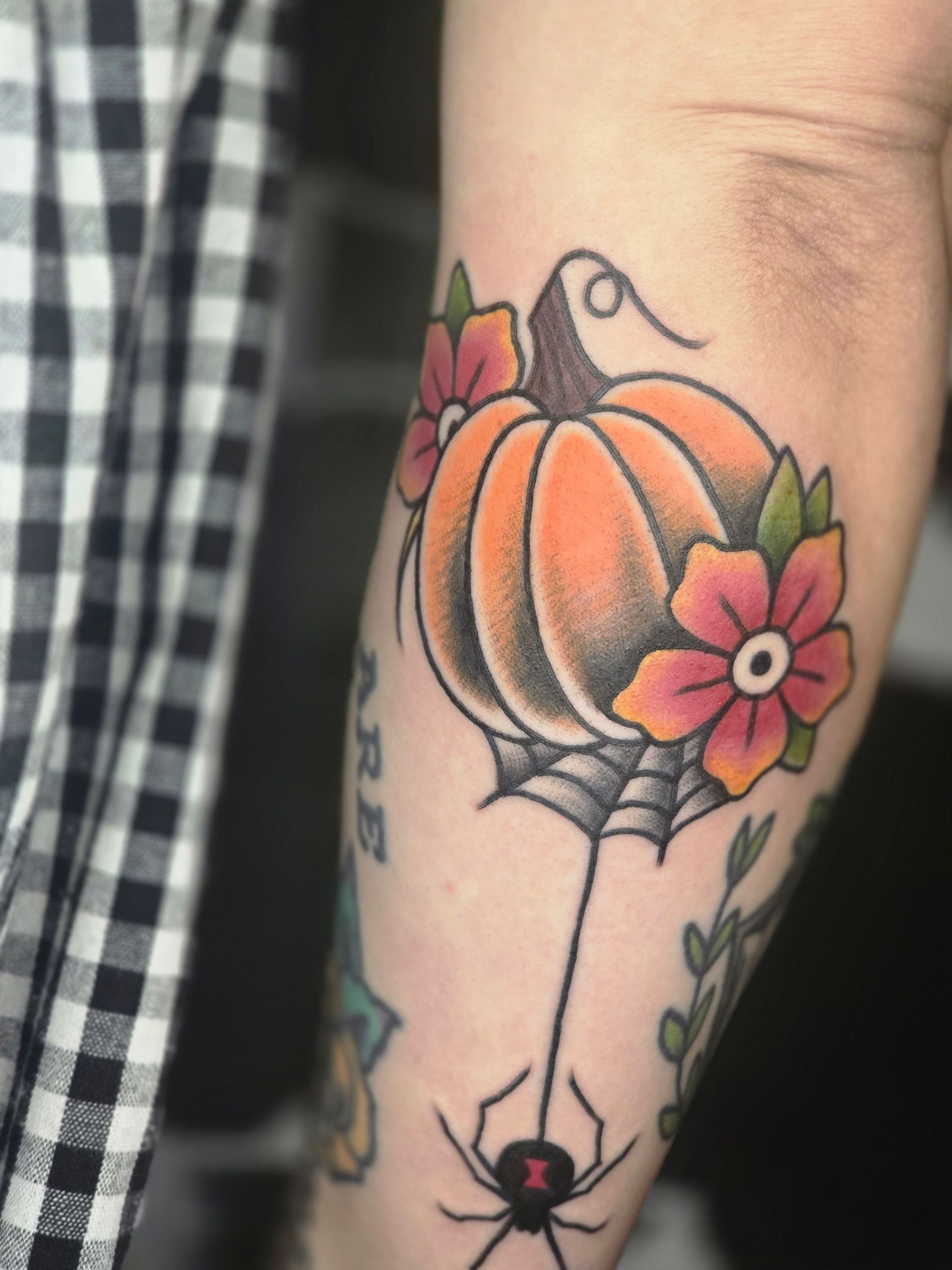 Tattoo of an orange pumpkin with flowers and a spider hanging from a web on an arm.