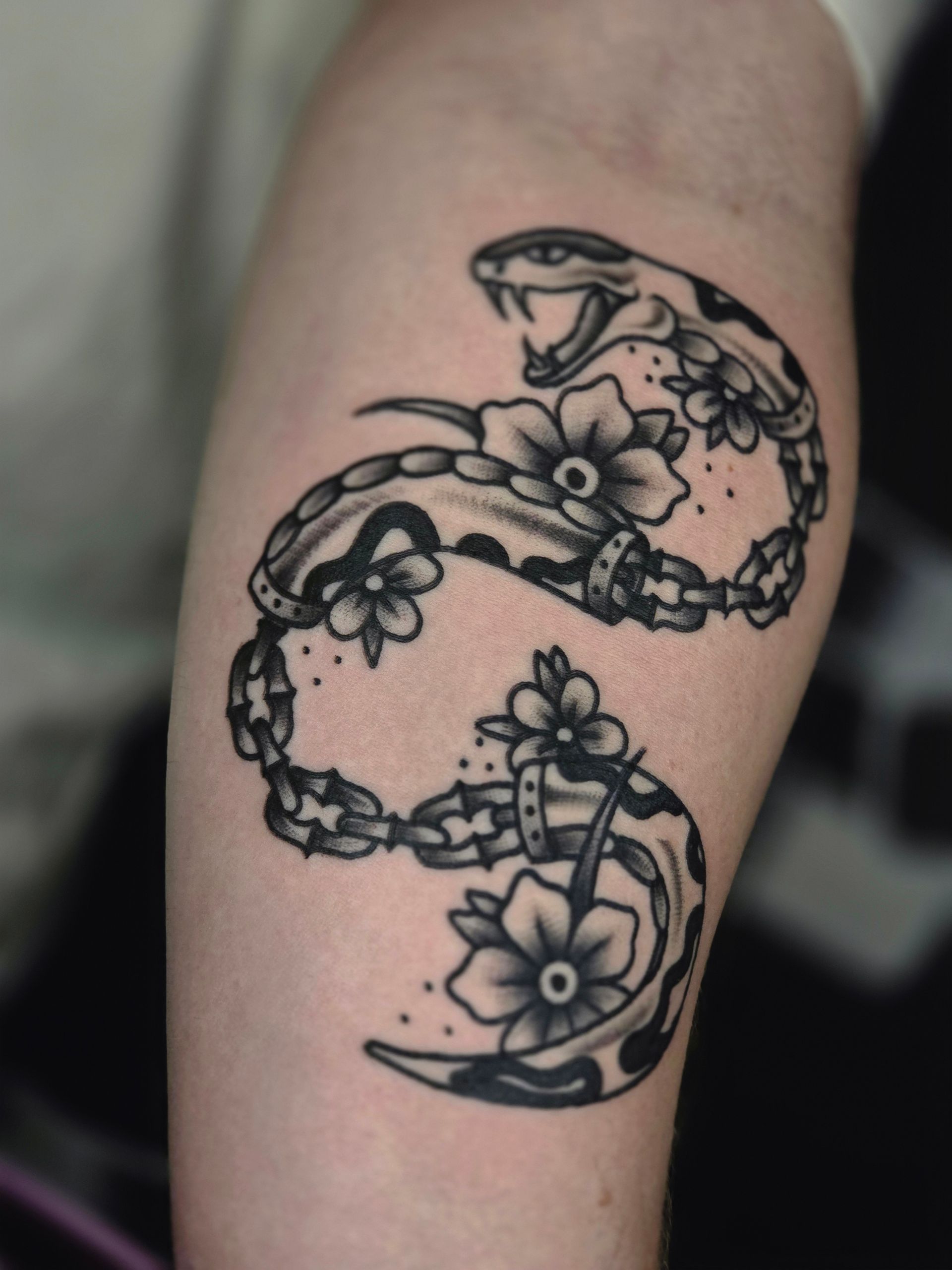 Black and white tattoo of a snake intertwined with flowers, snaking around an arm. Snake has open mouth.