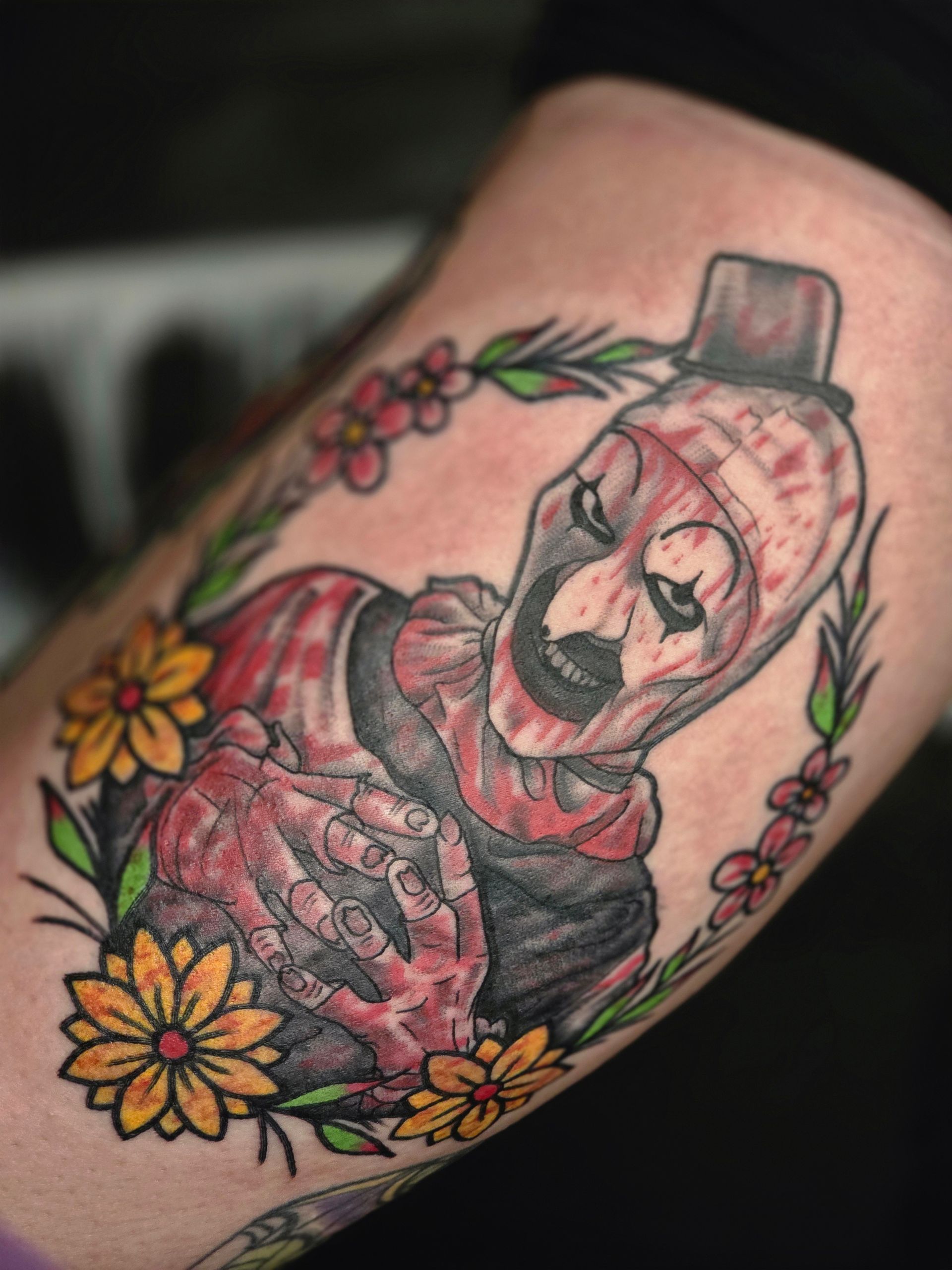 Tattoo of a bloody clown in a top hat, surrounded by a floral wreath, on a person's arm.