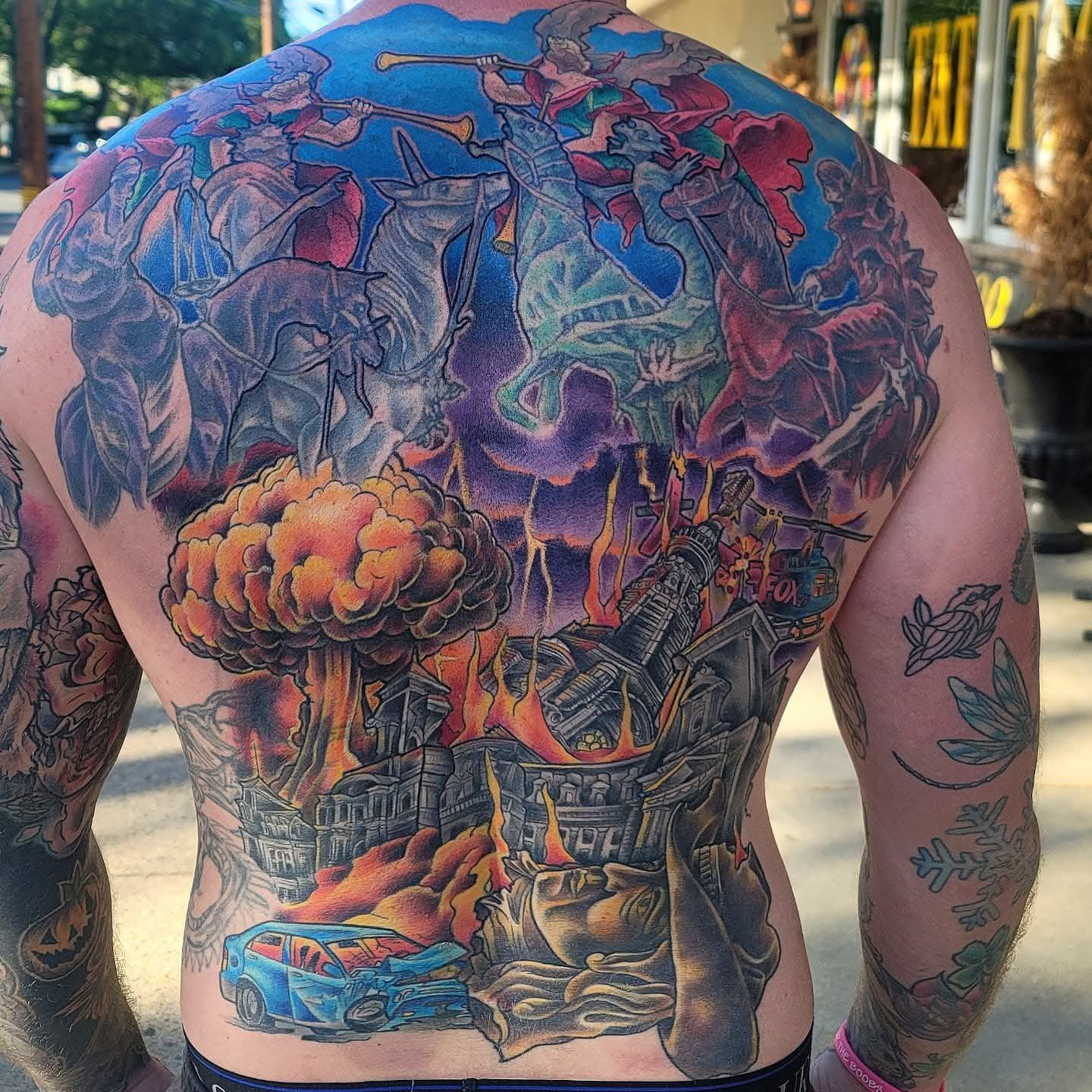 Back tattoo: a colorful, detailed depiction of an explosion, fire, and figures with a blue sky background.