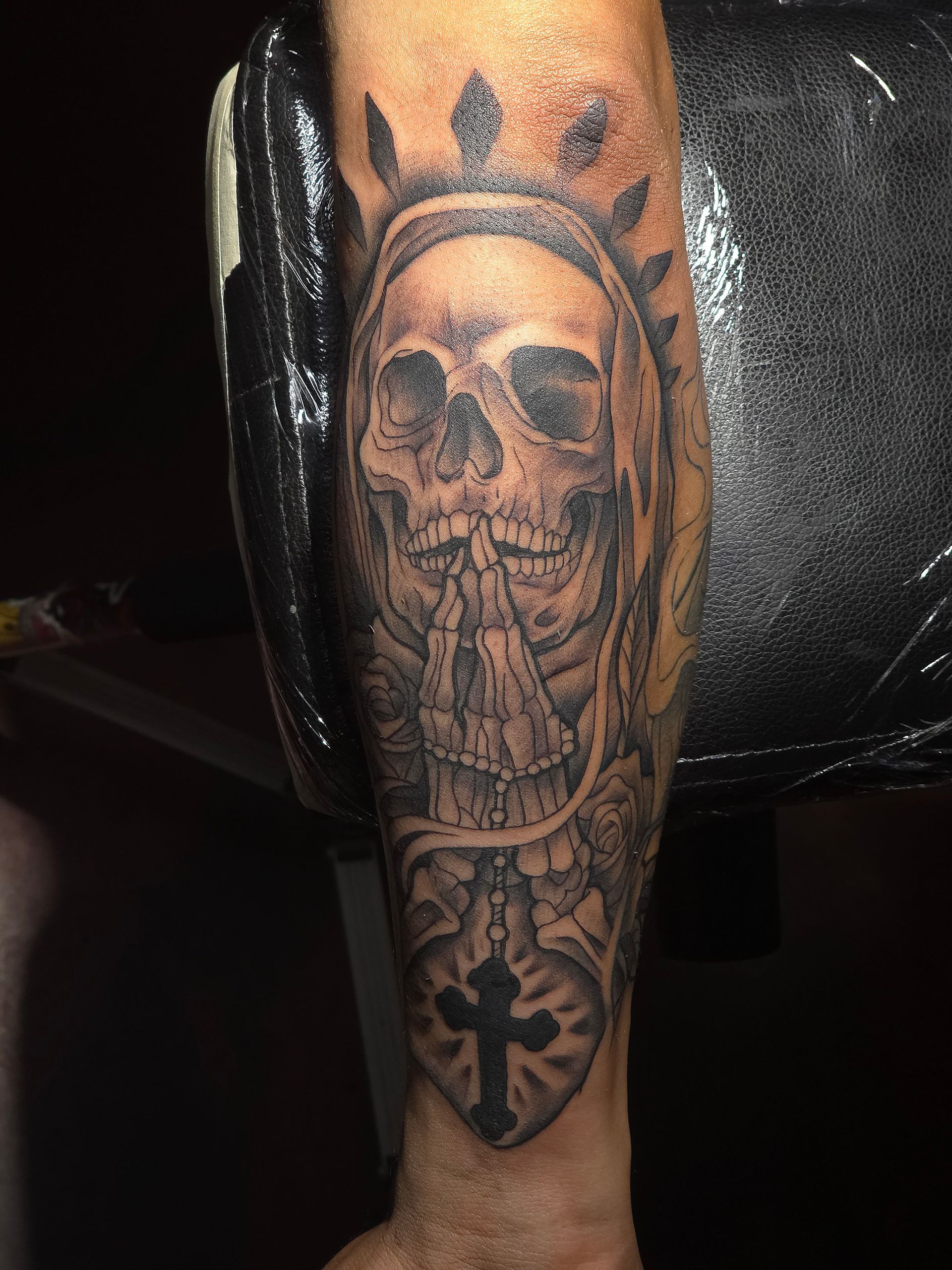 Tattoo of a skull with praying hands and a cross, on a person's arm. Black and gray shading.