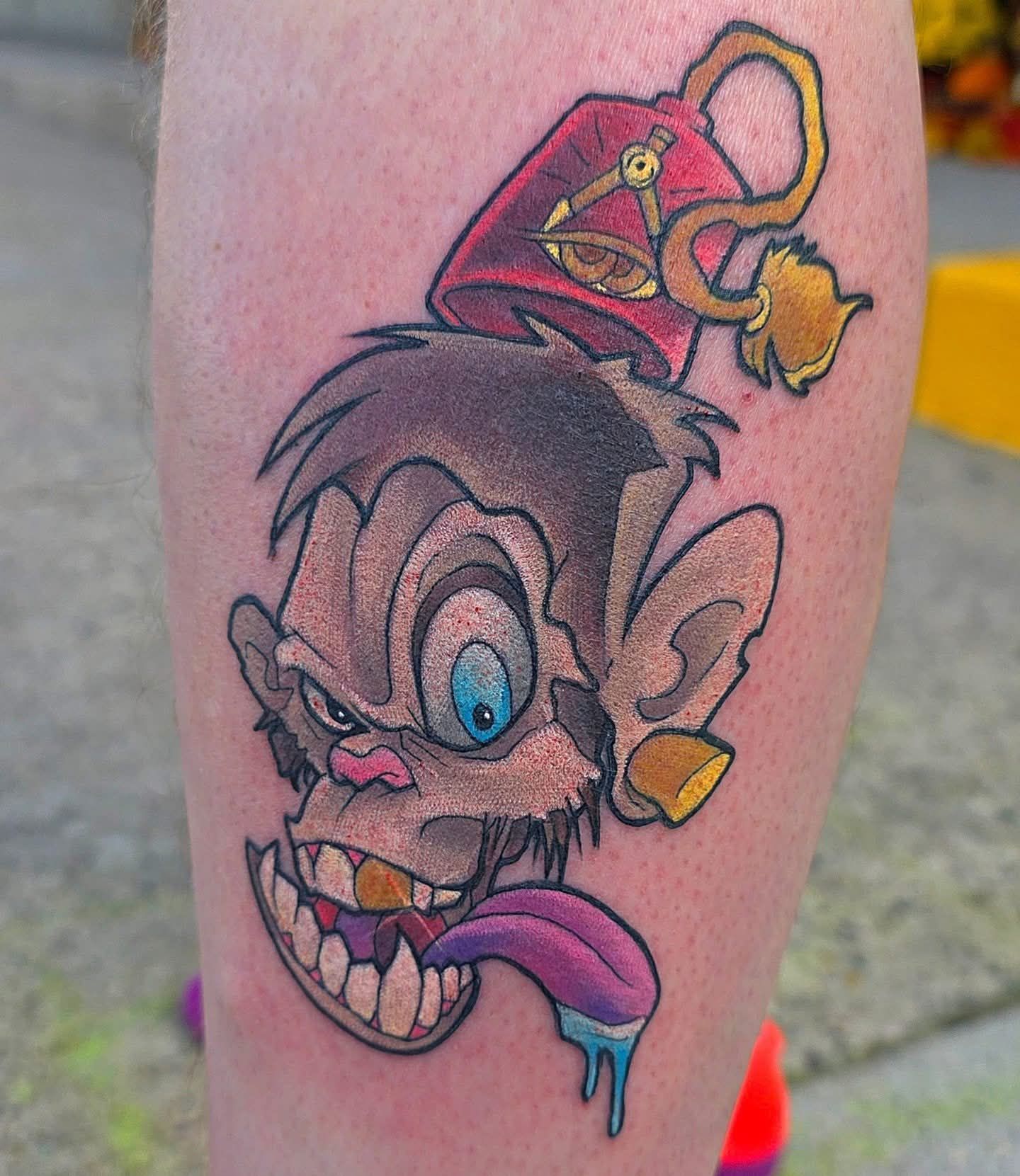 Tattoo of a cartoon monkey's face. It has bulging eyes, a long tongue, and wears a fez.