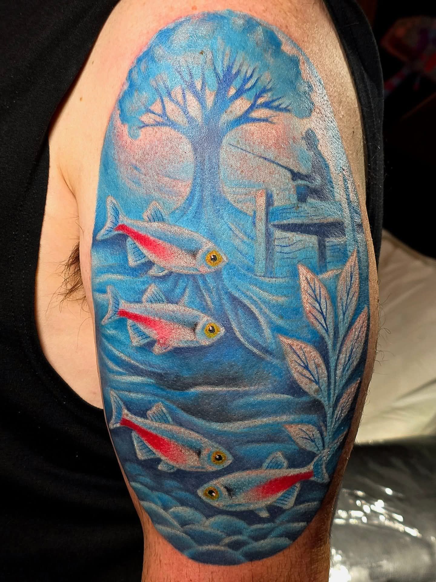 Tattoo of a fish swimming underwater with a tree, dock, and leaves in blue tones on a person's arm.