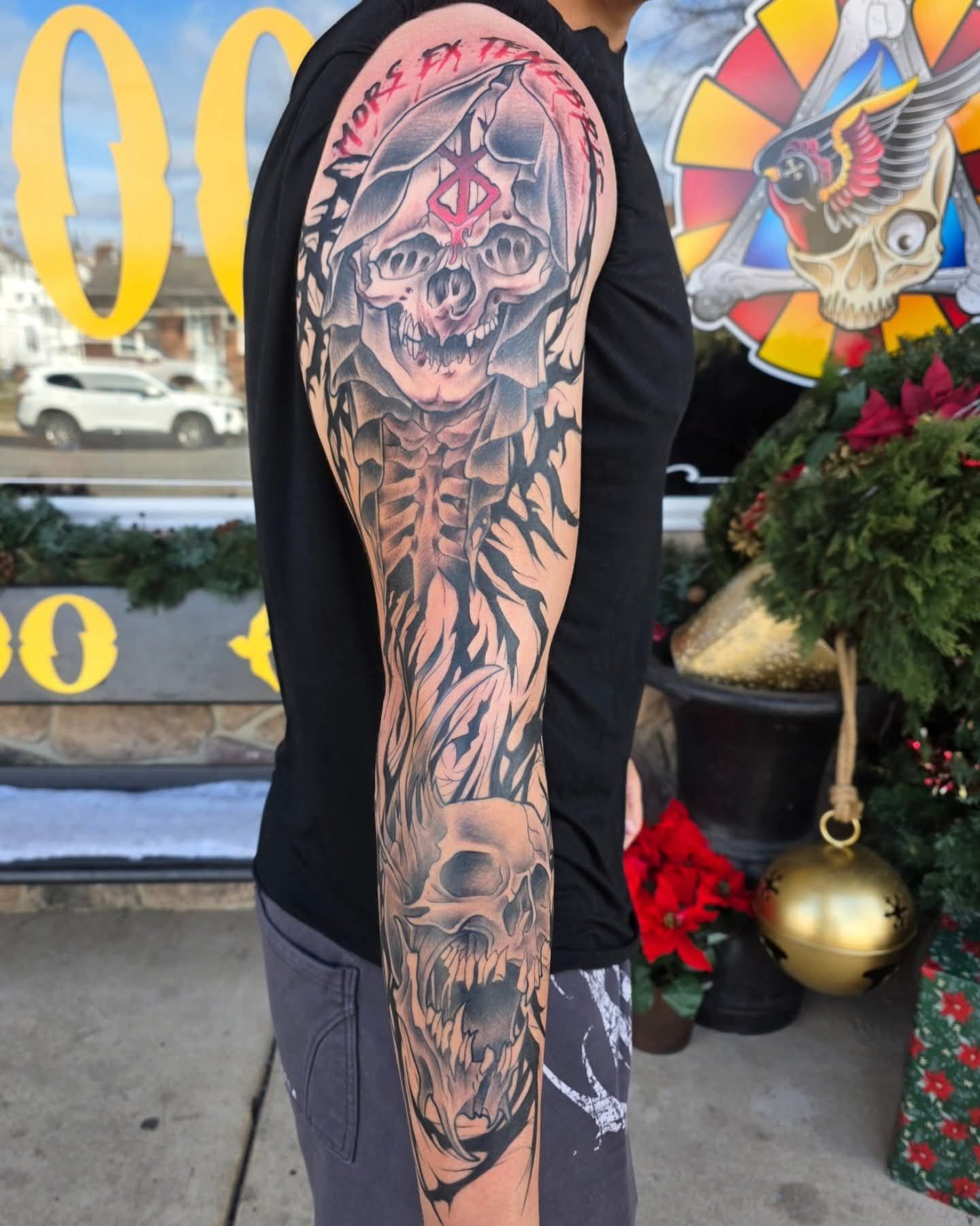 Man with full sleeve tattoo featuring a skull, skeleton, and stylized imagery. Outdoors by a shop.