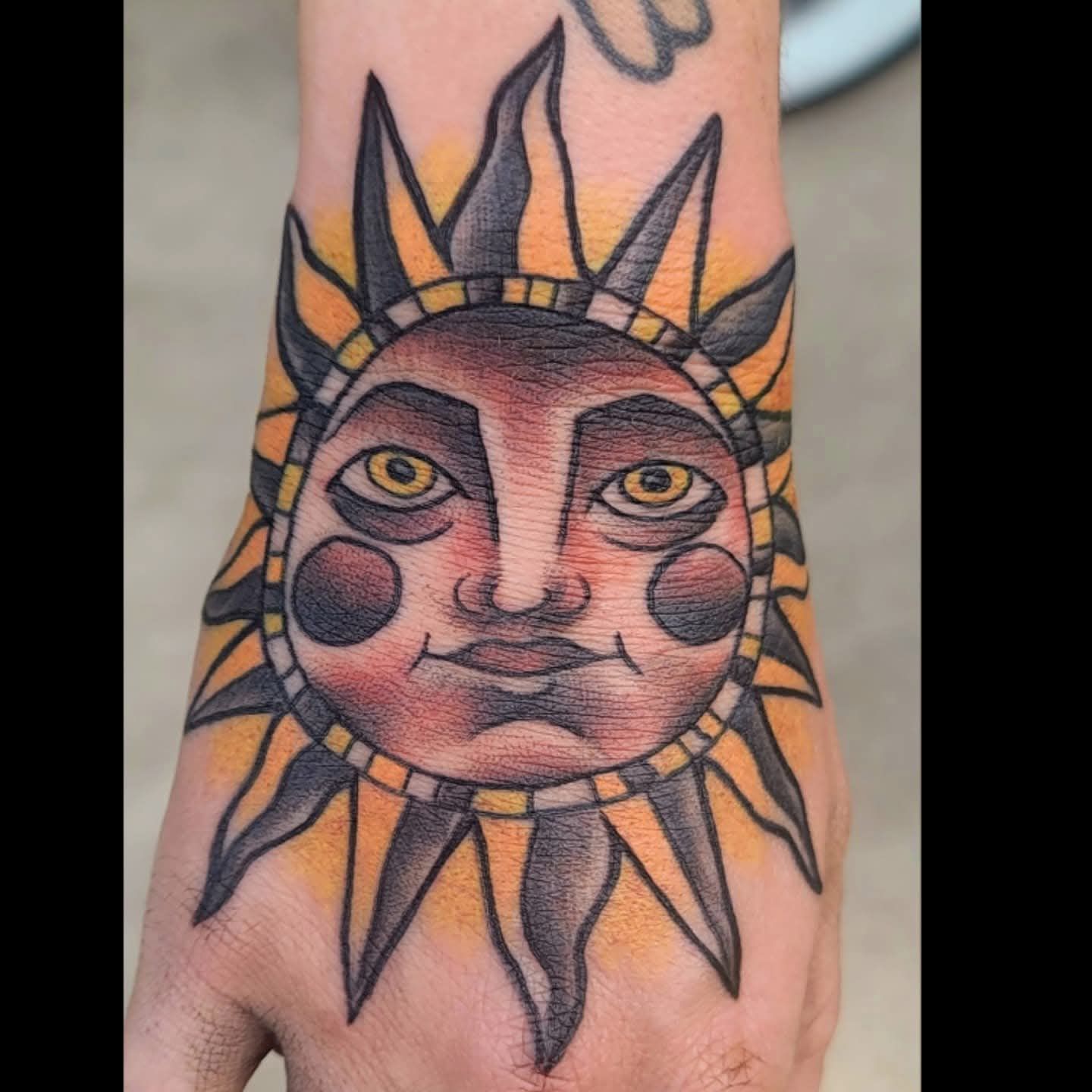 Sun-faced tattoo on the back of a hand, traditional style. Orange, yellow, and black.