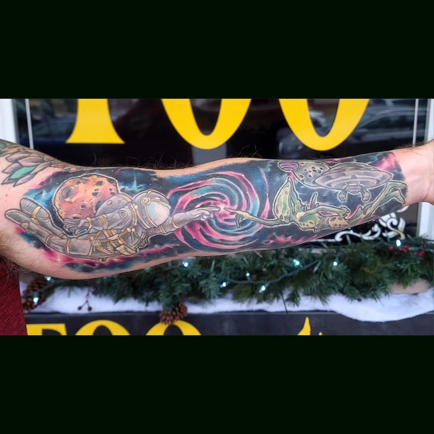 Arm tattoo depicting an astronaut reaching towards a cosmic swirl, connected to another figure with a halo, against a dark, starry background.