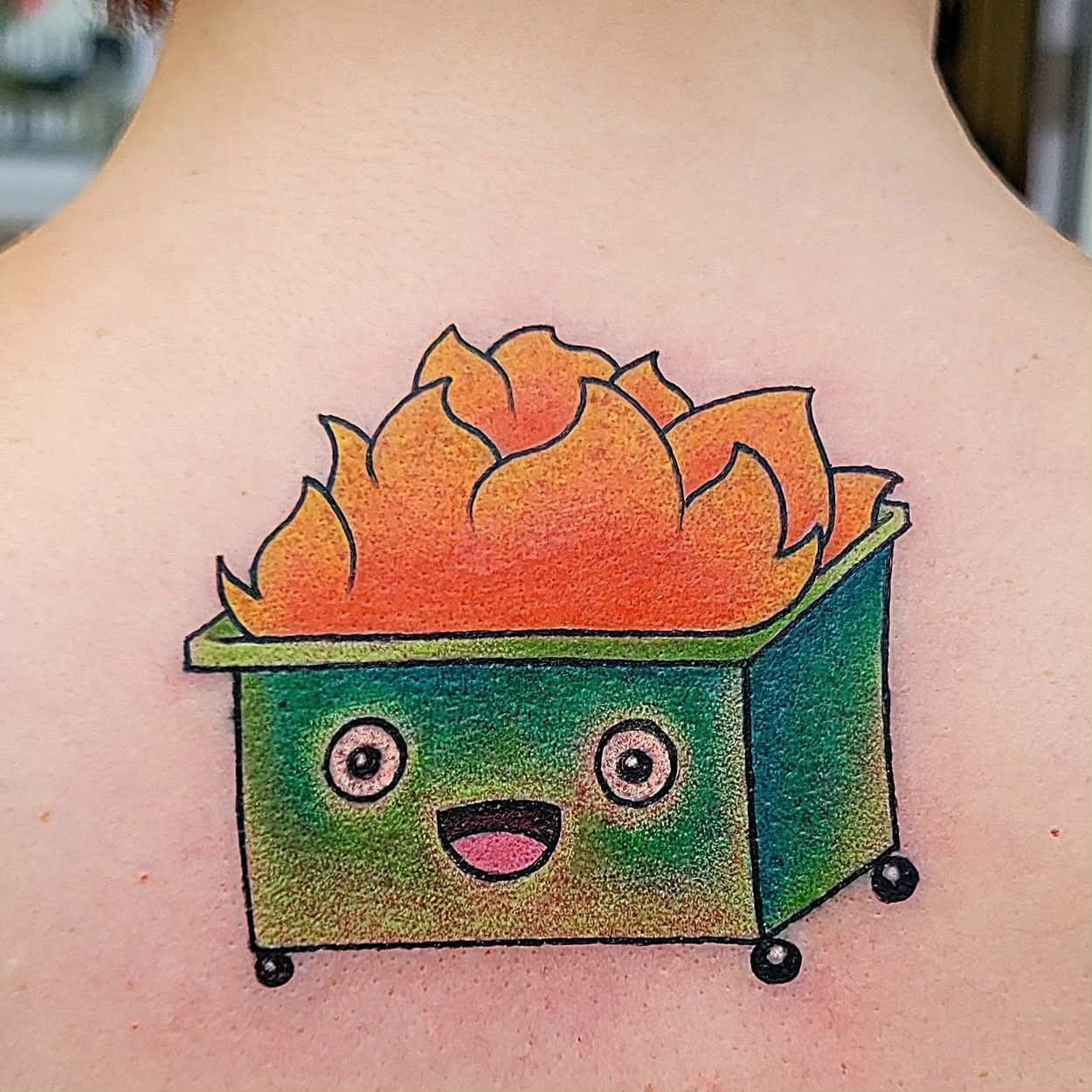 Tattoo of a smiling green dumpster on fire with orange flames, on a person’s back.