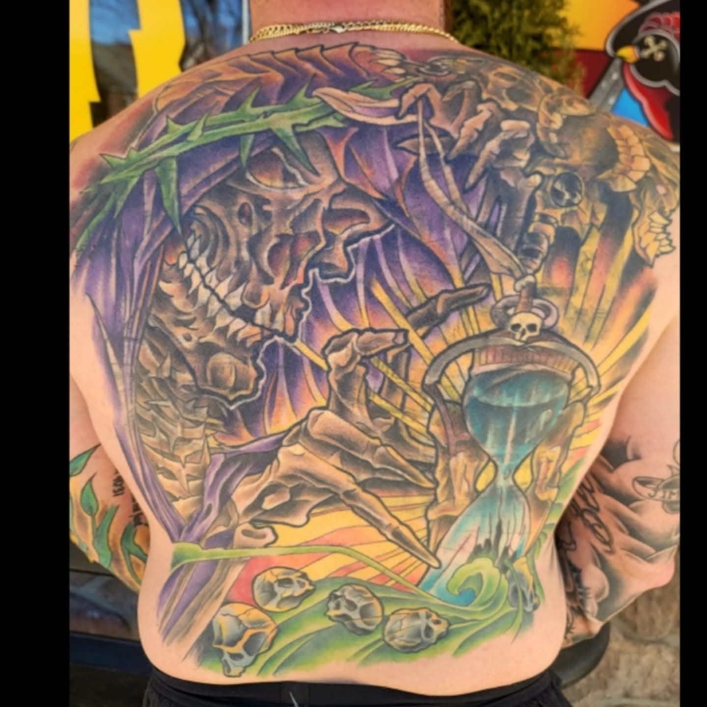 Back tattoo of a skeletal figure, an hourglass, and skulls in a surreal landscape with vibrant colors.