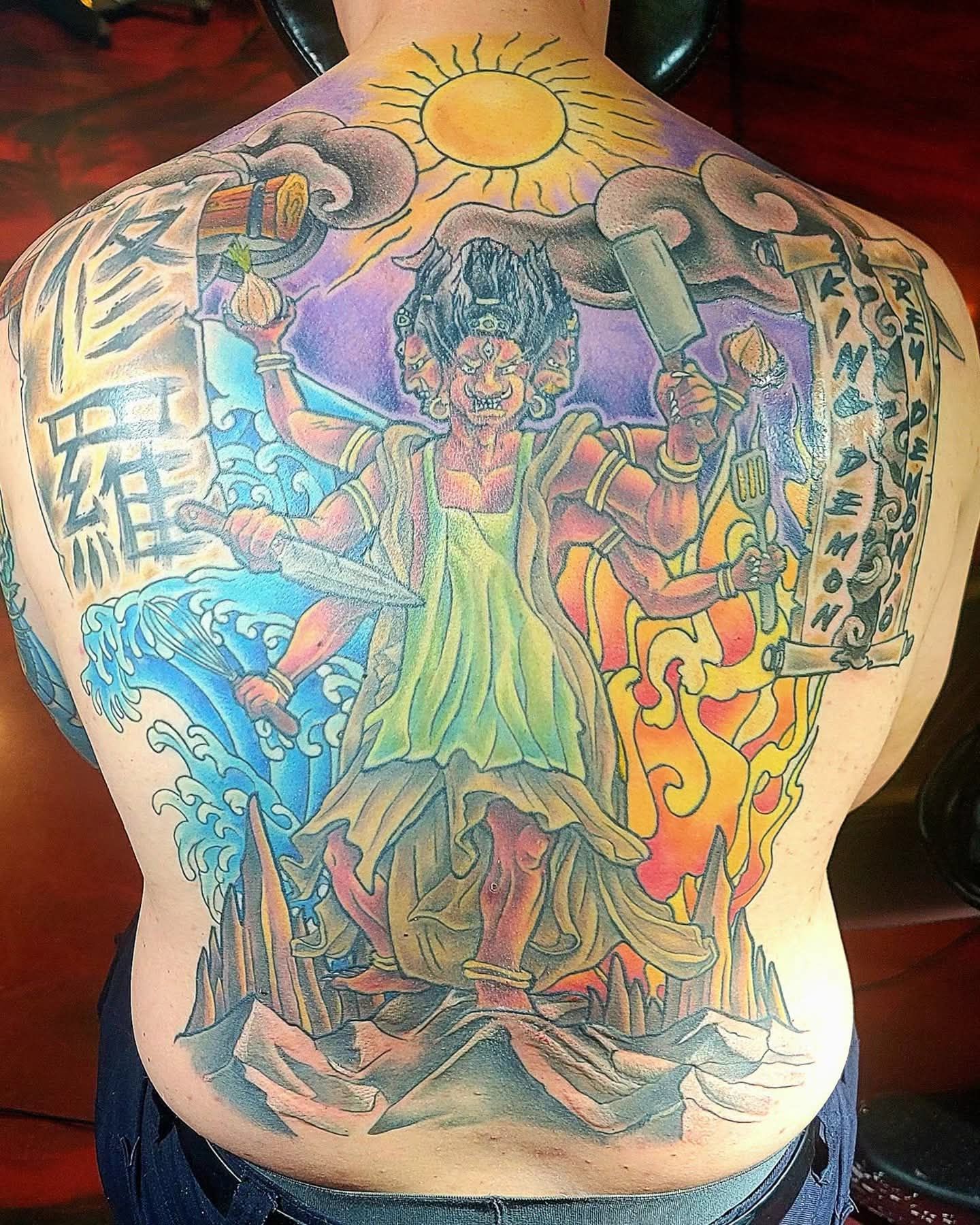 Back tattoo depicting a multi-armed deity standing on rock formations, surrounded by fire, water, clouds, and sun.