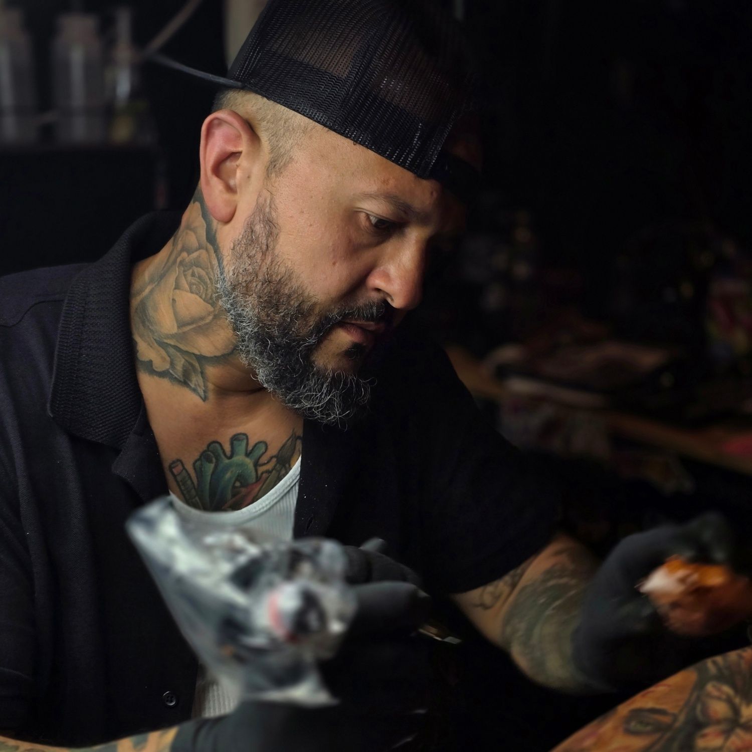 Tattoo artist in a dark studio, working on a tattoo. He has a beard, tattoos, and is wearing a black cap and gloves.