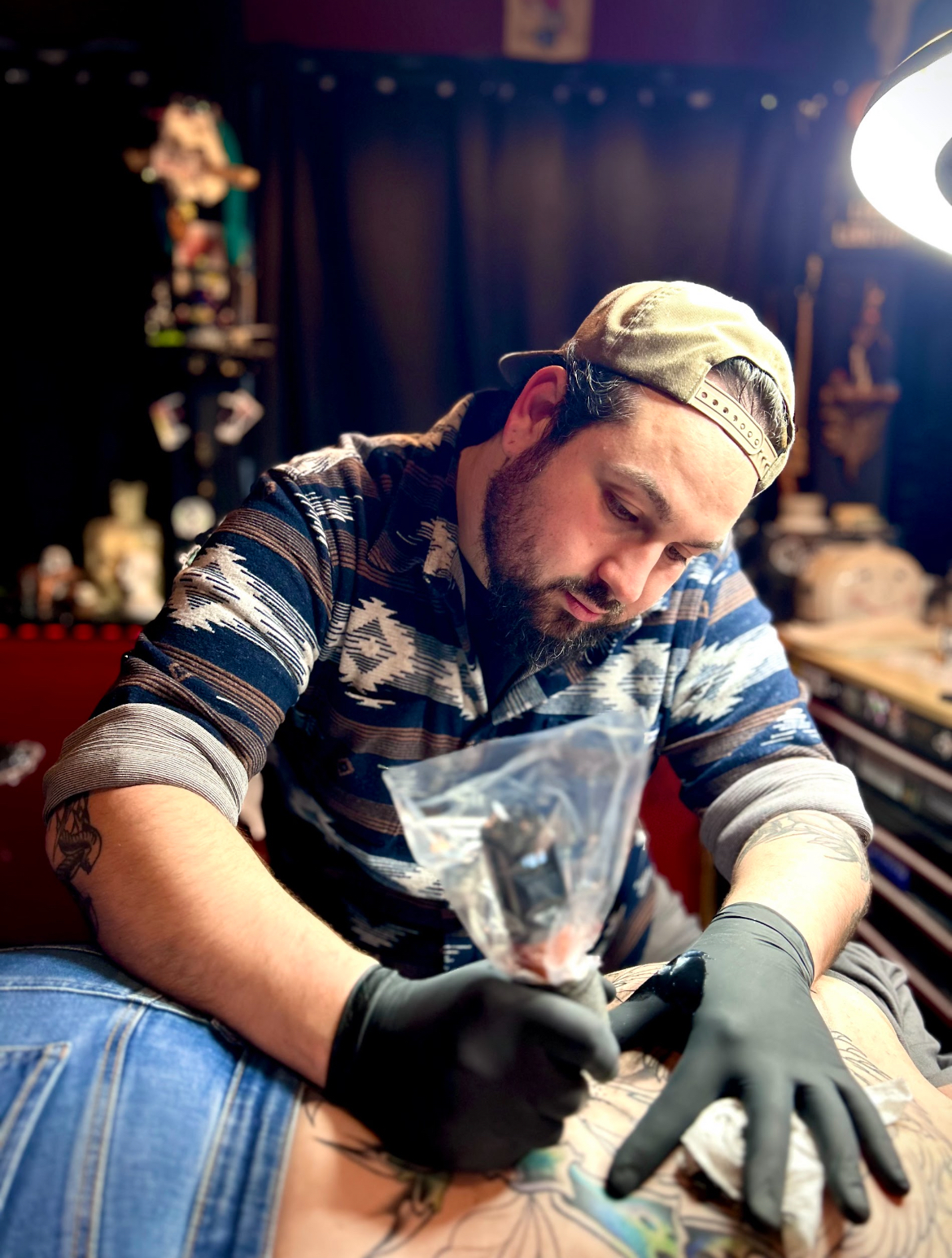 Tattoo artist in black gloves, using a machine on a person's back. Setting is a tattoo studio.
