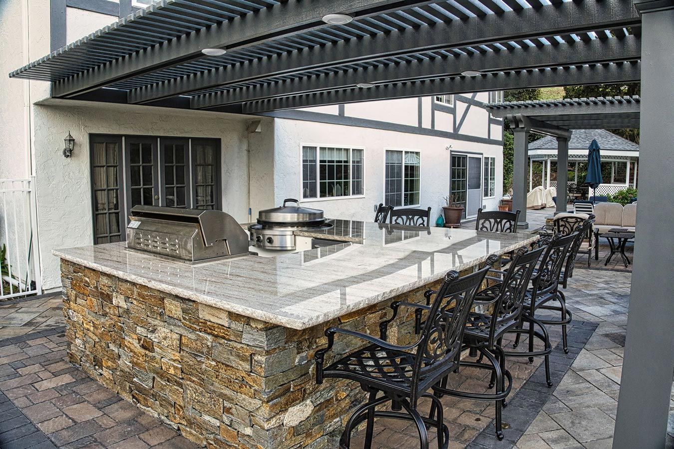 A large outdoor kitchen with a grill and chairs under a pergola.