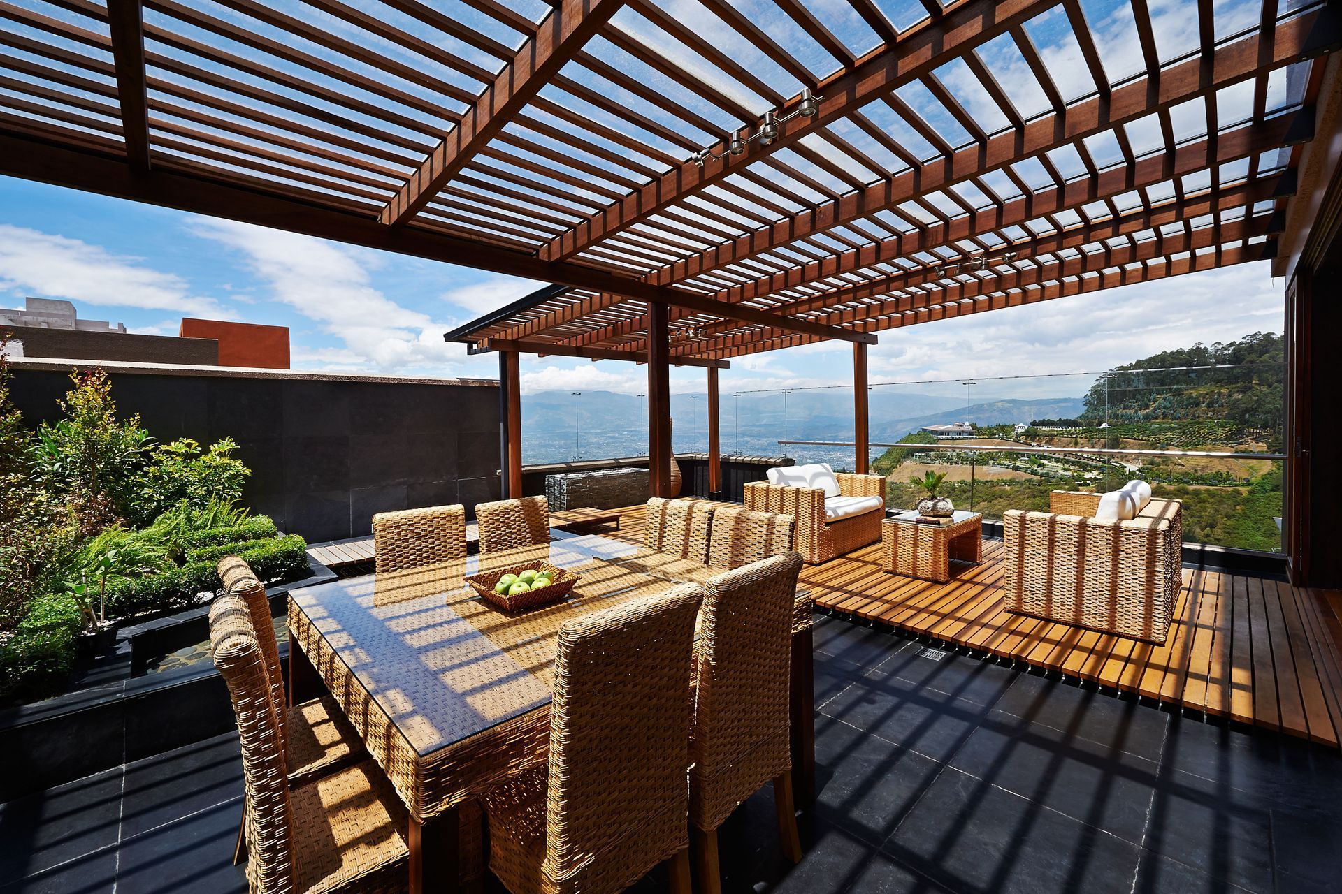 Outdoor patio dining area with wicker chairs, slatted pergola roof, and a scenic hillside view