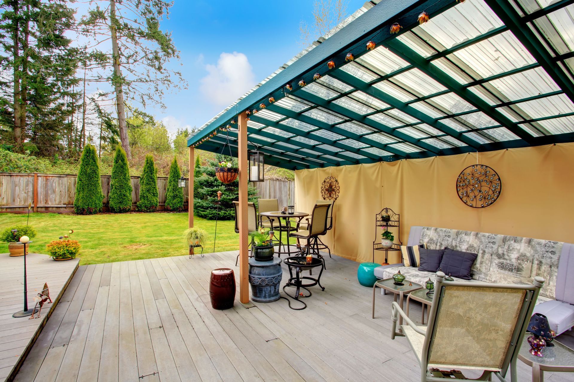 Covered backyard patio with wooden deck, seating, grill, and outdoor decor beside a grassy yard and trees