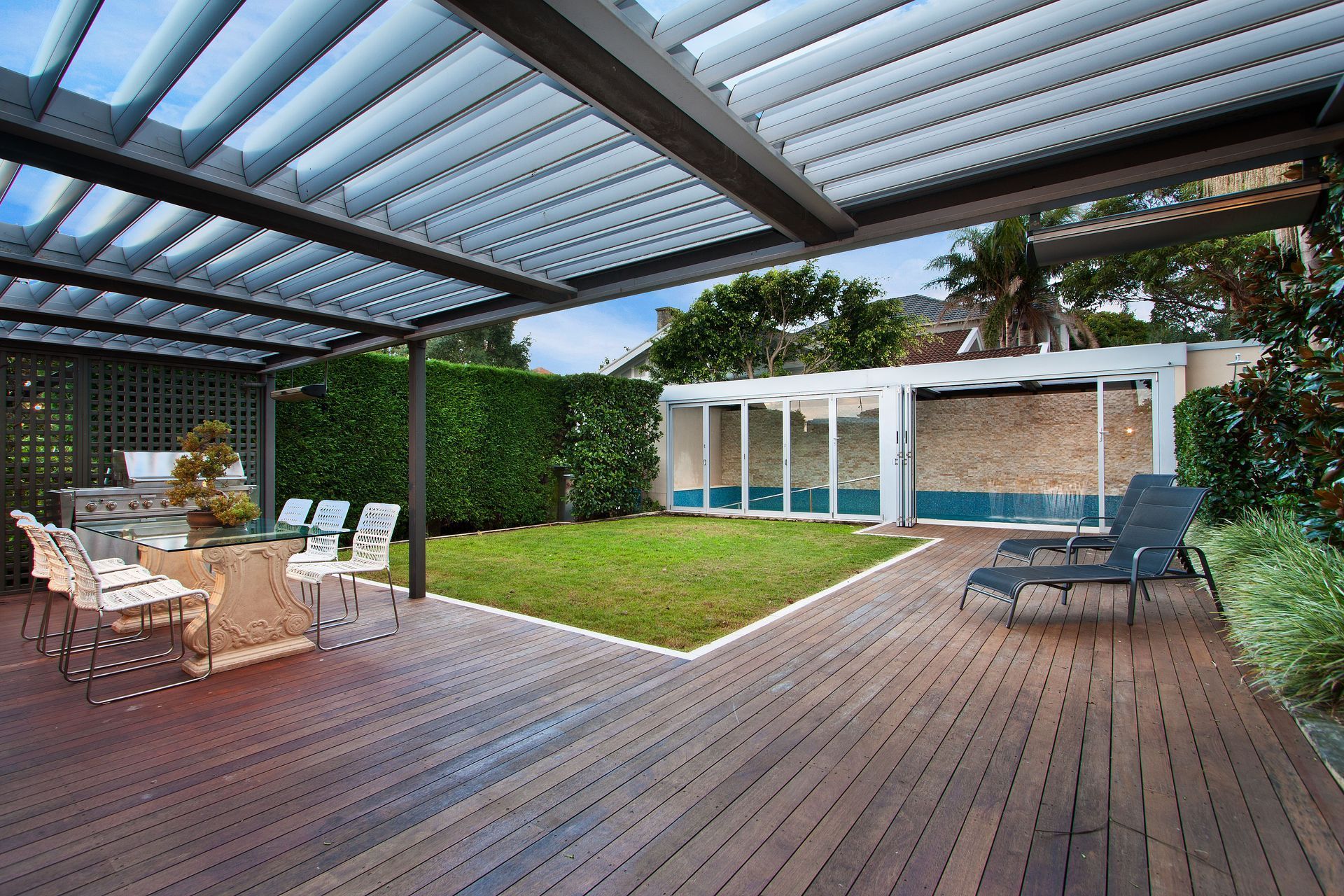 Covered wooden patio with dining table, lounge chairs, and a grassy yard beside a modern house.
