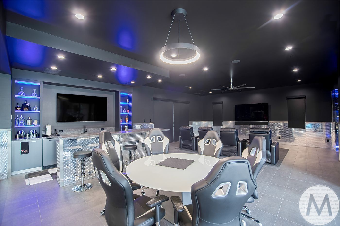 Modern lounge with black ceiling, round table, gray chairs, blue-lit shelves, and wall-mounted TVs