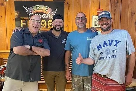 Four smiling people pose indoors in front of a wooden wall and sign, one giving a thumbs-up.