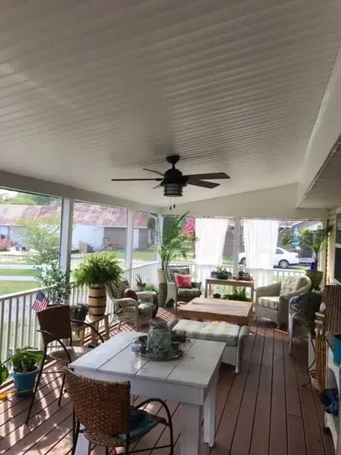 Covered porch with wicker seating, white dining table, ceiling fan, and potted plants overlooking a yard.